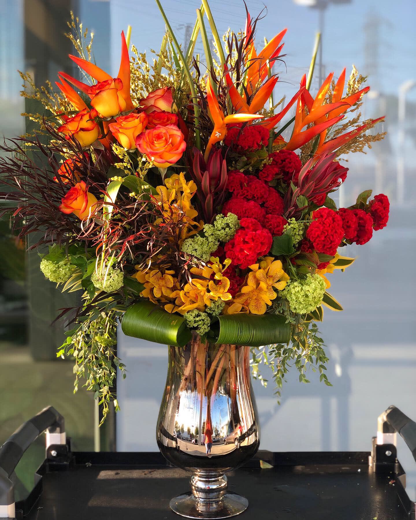 Tall bouquet of orange roses, red blooms, and orchids in a silver vase