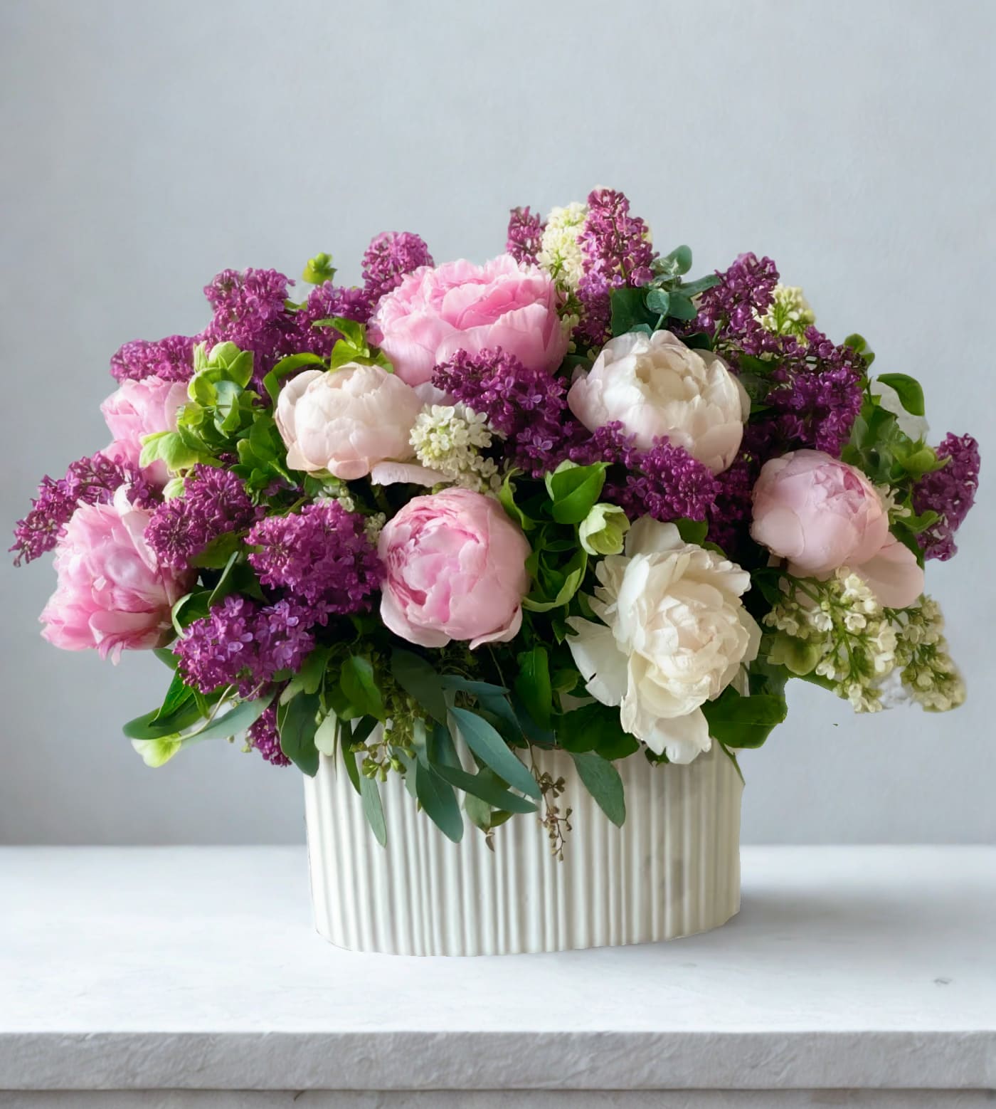 Pink and white peonies with purple lilac in a ribbed cream vase