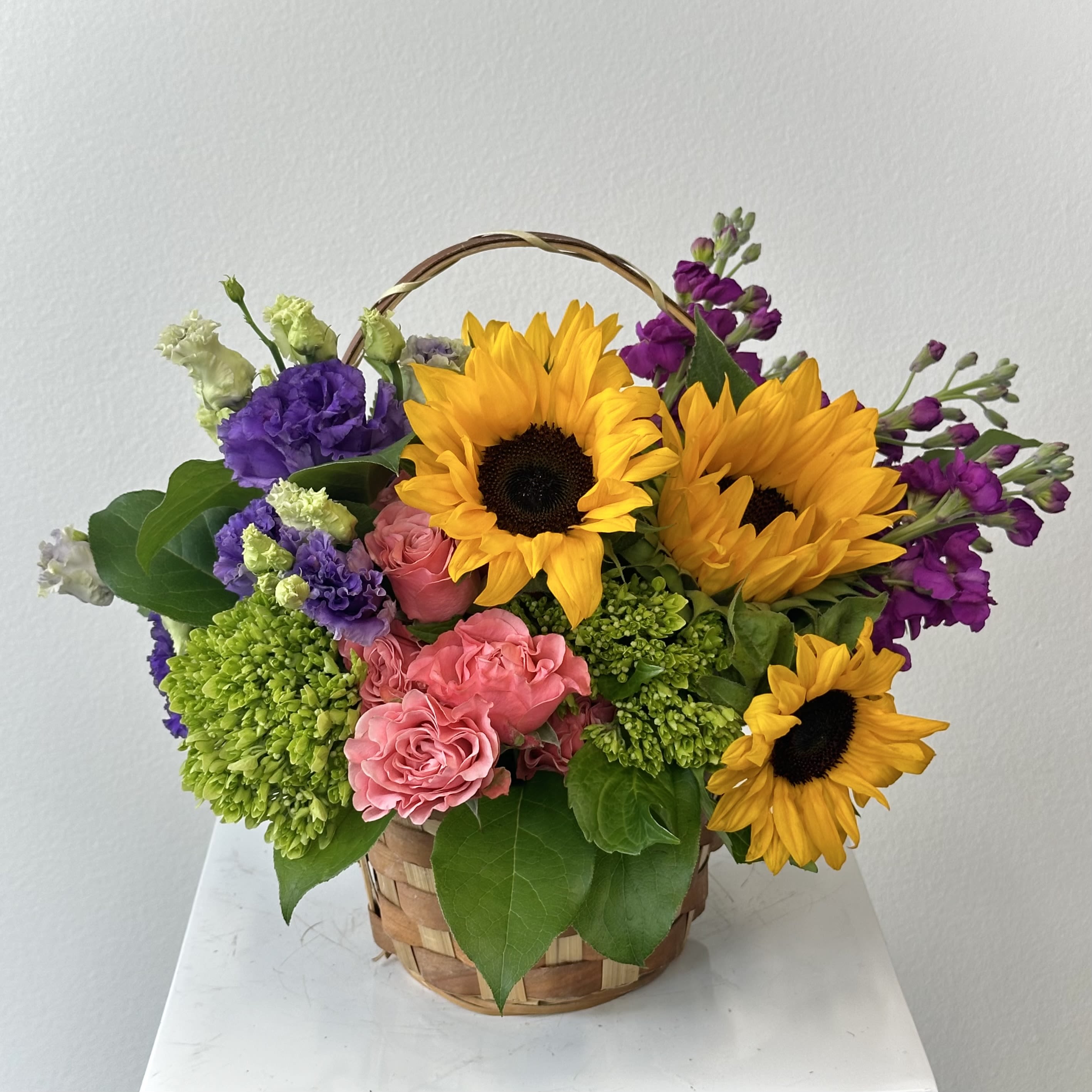 Picnic in the Park - A blooming delight for the garden-hearted! Bursting with sunshine and charm, the Picnic in the Park basket from Irvine Village Flowers is a miniature meadow crafted for your favorite blossom enthusiast—be it a green-thumbed granny or a floral fanatic. Picture this: towering yellow sunflowers stretching toward the light like summer’s golden rays, tender peach roses whispering romance and gratitude, regal purple stock standing tall with quiet elegance, petite green hydrangeas adding a lush, leafy hug, and spirited hot pink matsumoto asters dancing with playful energy. Nestled in a rustic rectangular handled basket, this bouquet is kissed by nature’s finest greens—sprightly solidago, wild huckleberry, and dappled variegated pittosporum—creating a lush, all-around wonder measuring approximately 12 1/2" W x 18" H.  Each flower tells its own tale: sunflowers beam with joy and adoration, peach roses bloom with warmth and appreciation, purple stock offers beauty and resilience, green hydrangeas bring heartfelt abundance, and hot pink asters sprinkle in a dash of cheer and charm. Perfect for brightening a kitchen nook, bedside perch, or coffee table haven, this arrangement is a heartfelt way to shower your loved one with nature’s love. Swing by Irvine Village Flowers online or in-store, and let us deliver this garden-fresh treasure straight to their door, blooming with joy!