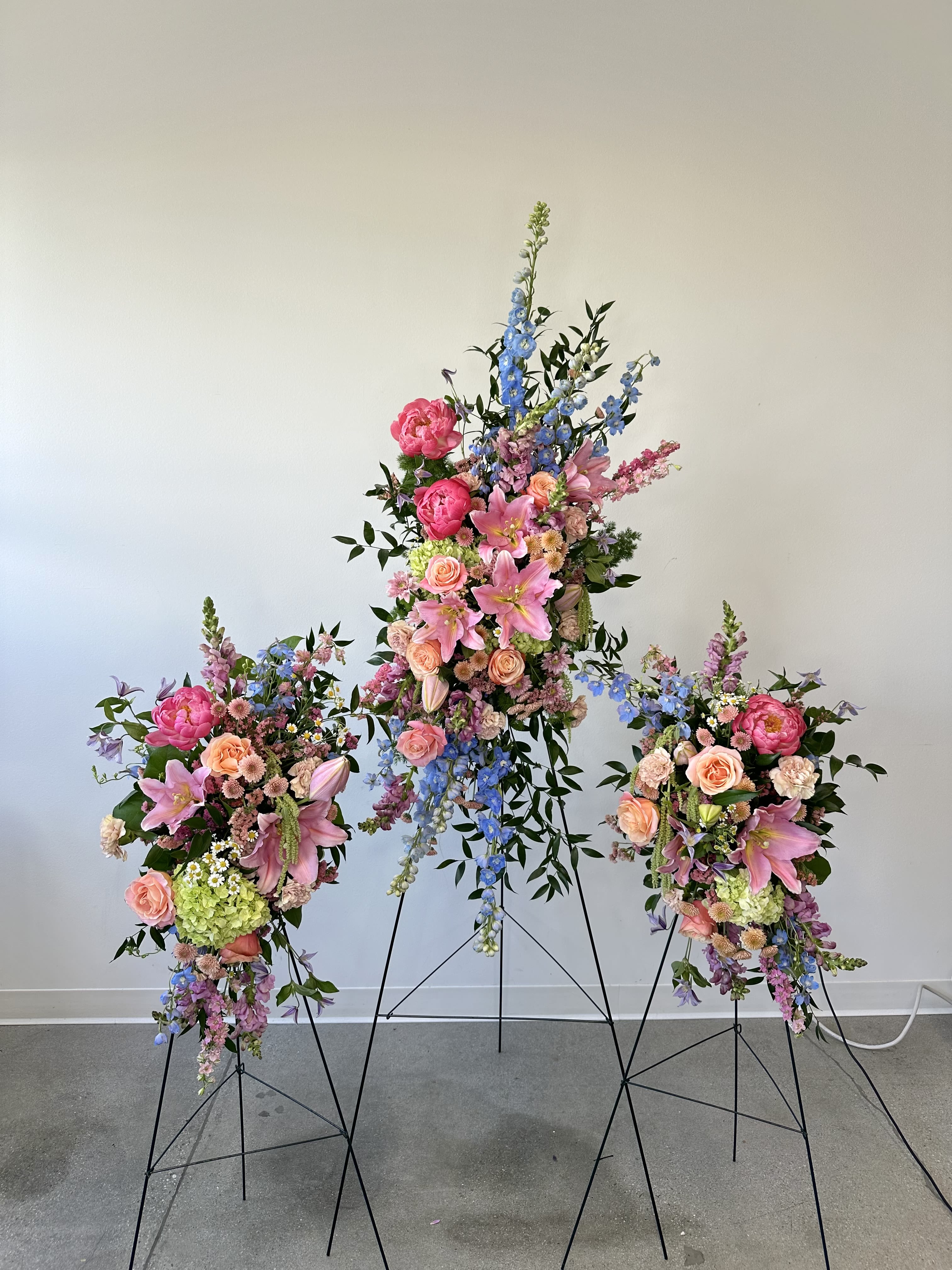 Eternal Garden Trio - Honor a cherished life with the Eternal Garden Trio, an extraordinary sympathy floral spray set featuring one grand standing spray accompanied by two coordinating companion sprays. Designed to create a breathtaking and elegant memorial display, this garden-inspired tribute surrounds loved ones with beauty, comfort, and remembrance during funeral services, celebration of life ceremonies, and memorial gatherings. Each spray is handcrafted with premium fresh flowers, including fragrant pink lilies, luxurious pink peonies, vibrant blue delphinium, lush green hydrangeas, peach and pink roses, graceful lavender snapdragons, cascading green hanging amaranthus, soft peach carnations, and delicate peach and lavender chrysanthemums.  The trio arrangement creates a stunning visual presence, with the large central spray serving as the focal point while the two smaller companion sprays provide balance and fullness. The harmonious blend of pink, peach, lavender, blue, and green tones evokes the peaceful beauty of a flourishing garden, symbolizing love, admiration, hope, and everlasting memories. Flowing textures, premium blooms, and thoughtful design make the Eternal Garden Trio a meaningful expression of sympathy and support for family and friends. Whether displayed at a funeral home, church, memorial chapel, or graveside service, this elegant three-piece floral tribute offers a heartfelt way to celebrate a life well lived while providing comfort and encouragement to those left behind.