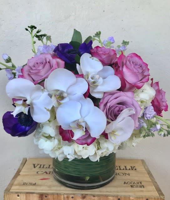 Round arrangement of white orchids, hydrangeas, and pink and purple roses in a glass vase.