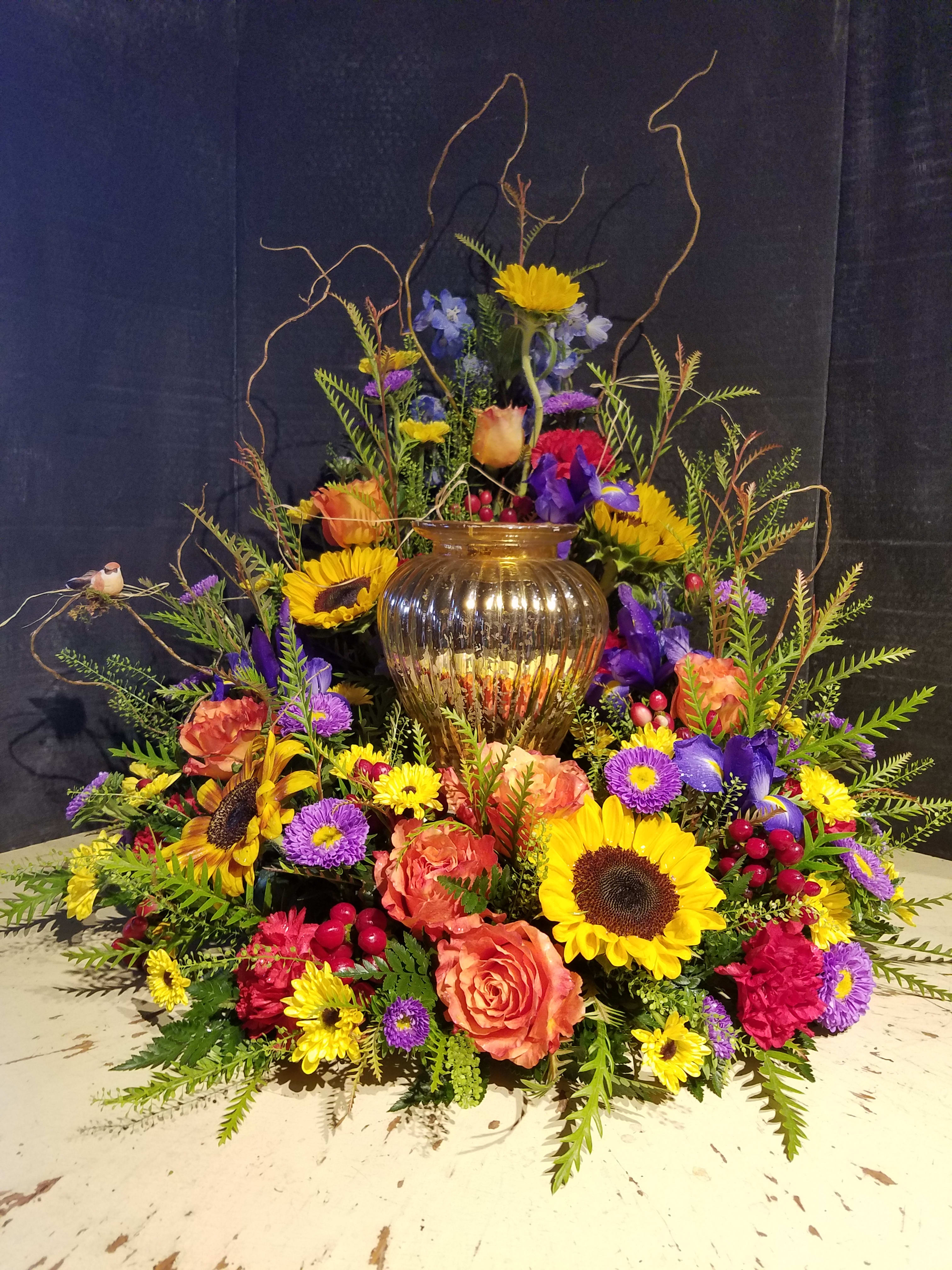 Large mixed floral arrangement with sunflowers, roses, and purple blooms in a gold vase