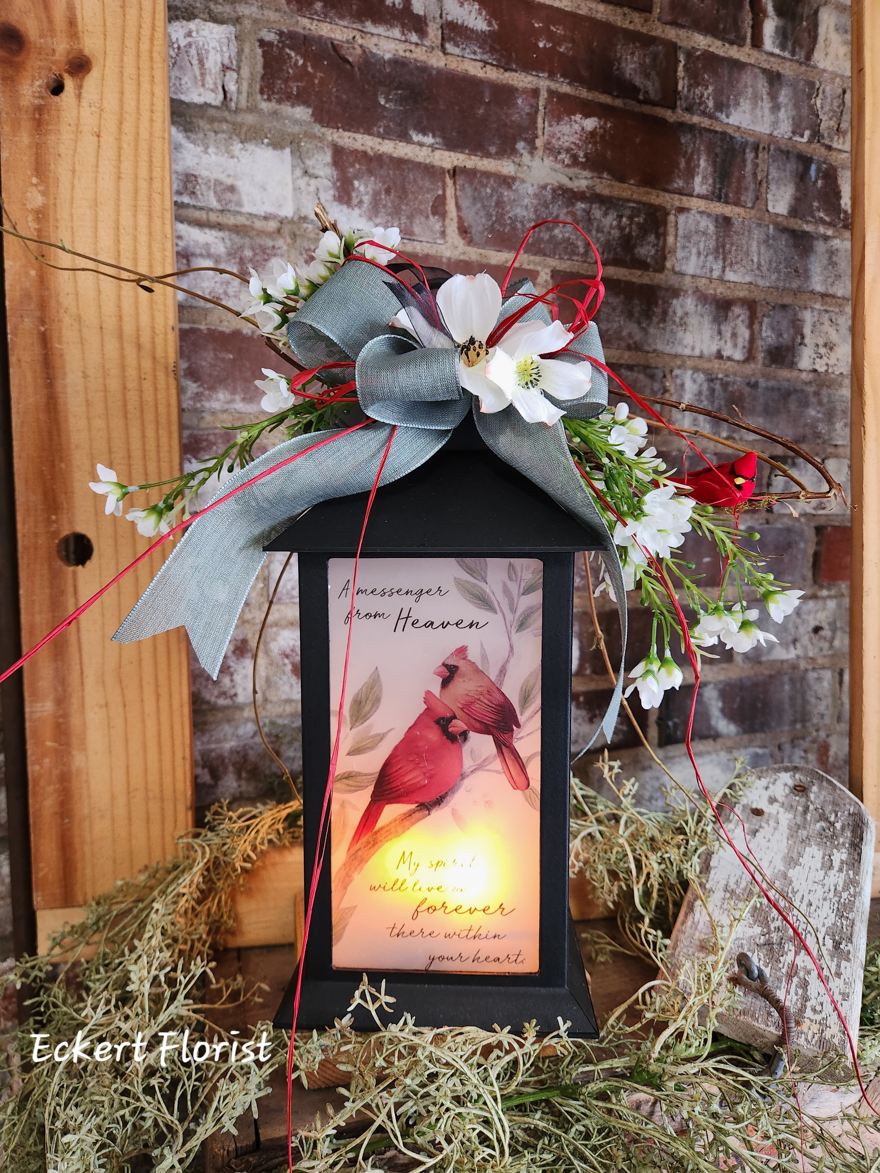 Eckert Florist's "A Messenger from Heaven..." Cardinal Lantern *Our Local delivery Only - This realistic battery-operated flickering LED flame Lantern made with a high quality polyresin finish, decorated with silk flowers, designer ribbon, and our signature bird, is a great memorial gift. The message printed on the frosted glass reads: "A messenger from Heaven - My spirit will live on forever there within your heart." Lantern measures approx. 13" H x 5.5" W x 5.5" D. *Batteries included *Our Local Delivery Only