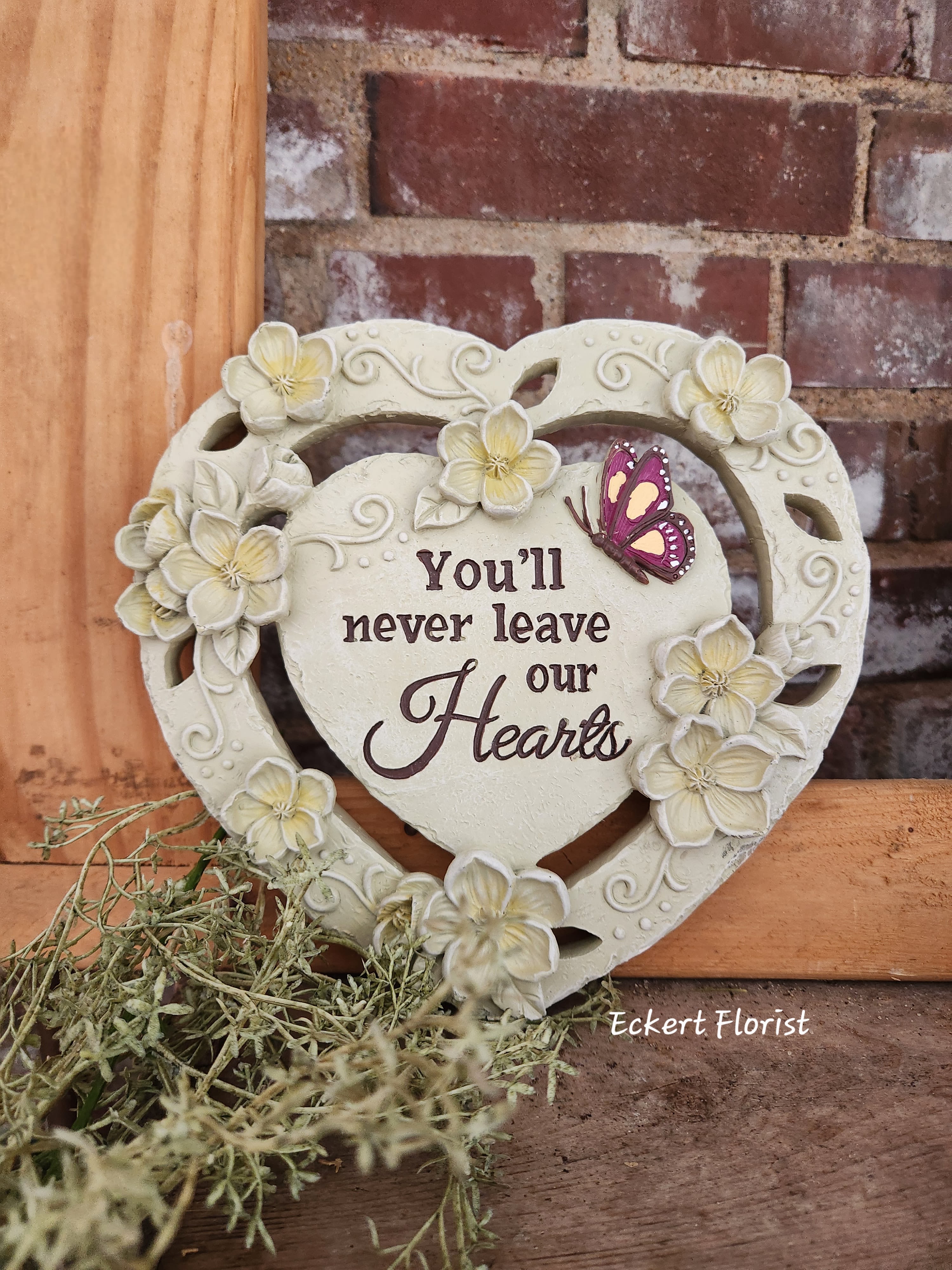 Heart-shaped decorative plaque with flowers and a butterfly, reading "You'll never leave our Hearts"