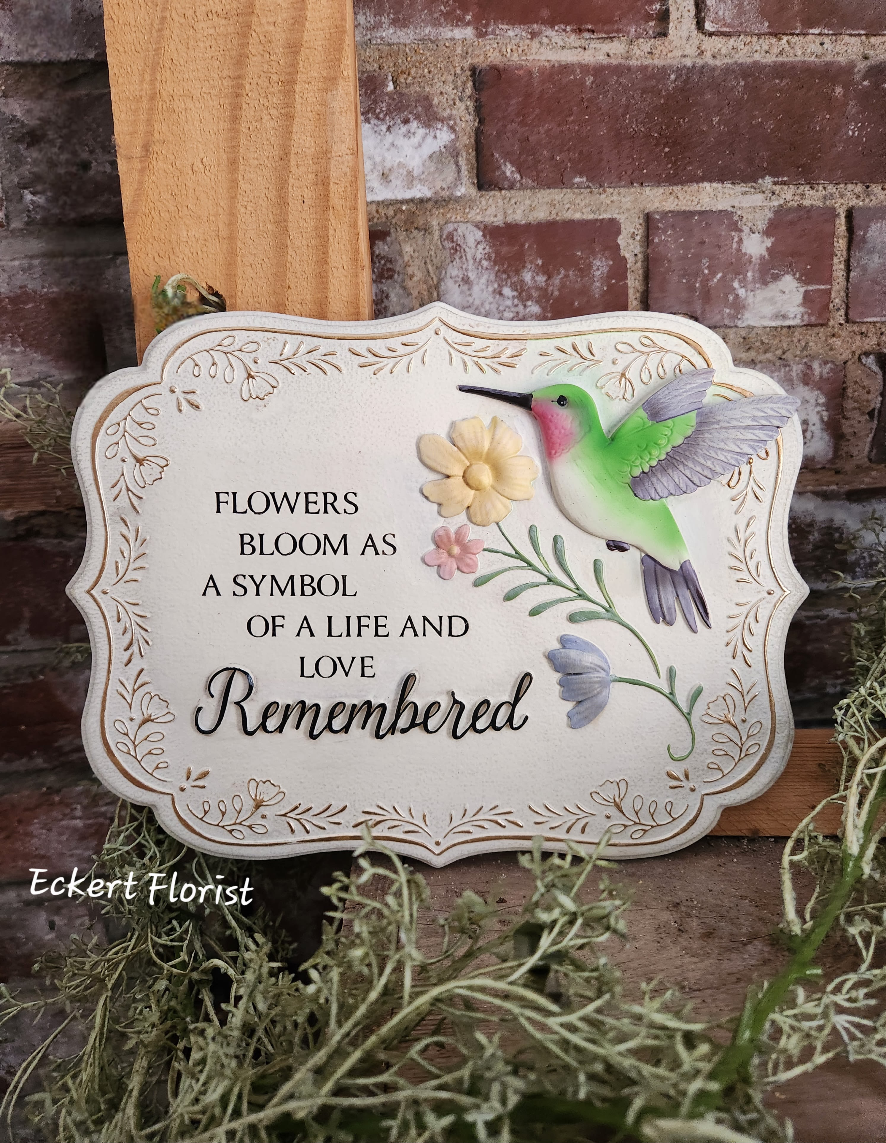 Eckert Florist's "Remembered" Garden Memorial Stone - This beautifully crafted garden stone is made of hand-painted resin and adorned with a vibrant floral design and delicate hummingbird. These bright colors and charming details bring warmth and inspiration to any garden or outdoor space. W: 10 1/2" H: 8" D: 1/2 "