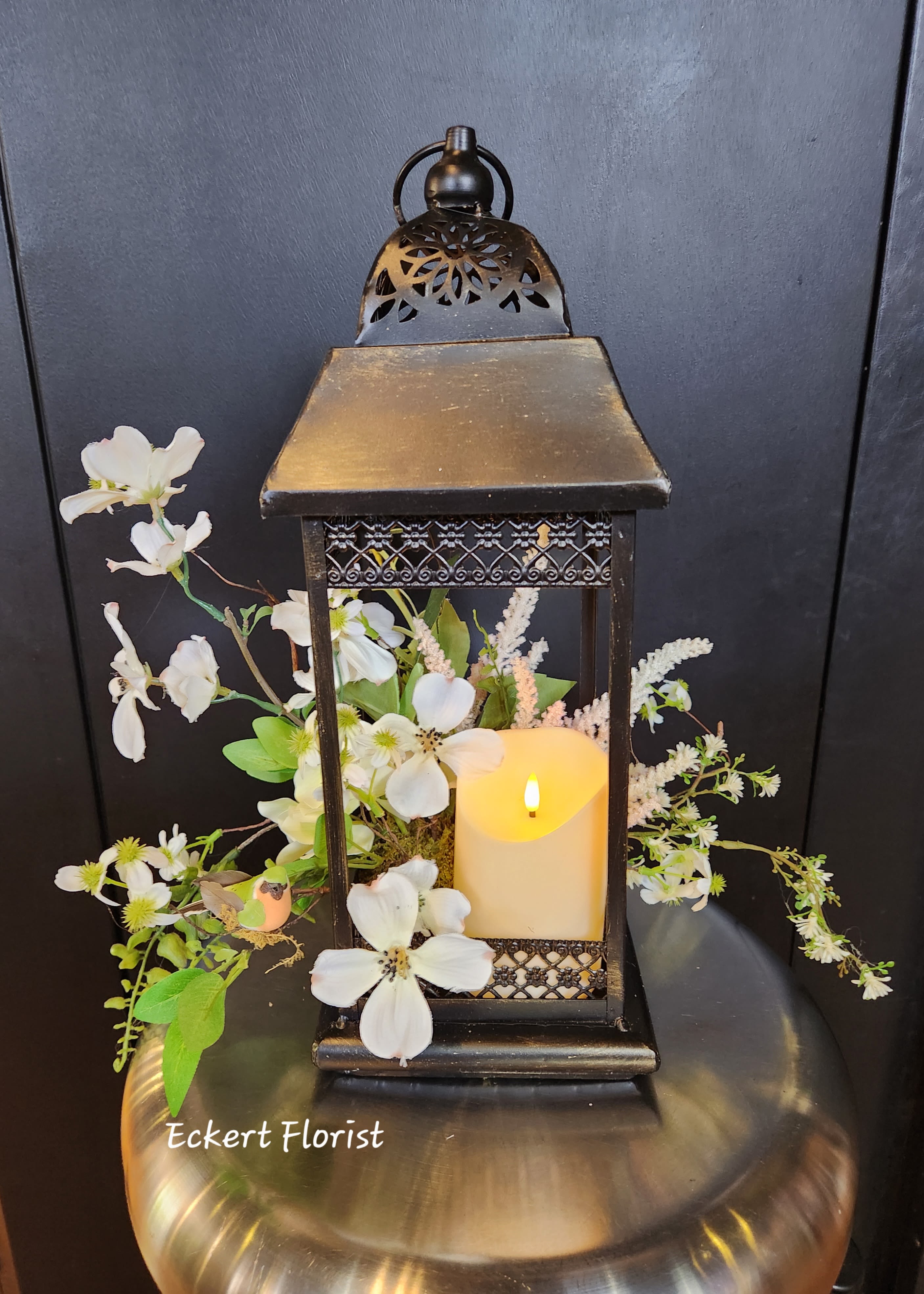 Eckert Florist's Bronze Metal Open Lantern - This bronze colored metal lantern is filled with a mix of white artificial flowers. Also included is a  battery operated candle to illuminate and inhance this eye catching design perfect for a memorial or any in-house decor. Lantern measures approx. 16" High 6" Wide 6" Deep (Batteries are included)