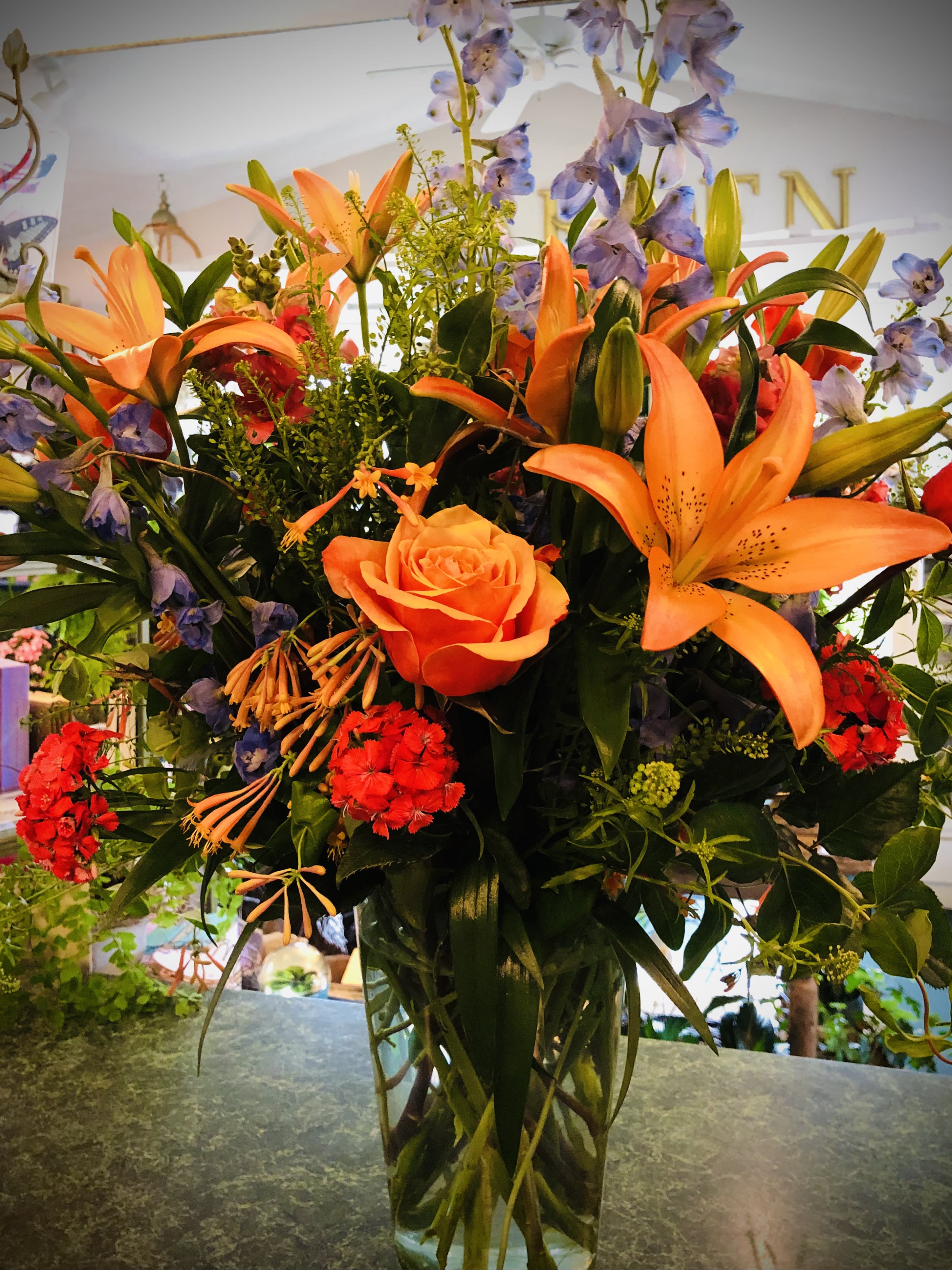Tall mixed arrangement with orange lilies, an orange rose, red blooms and blue delphinium in a clear glass vase