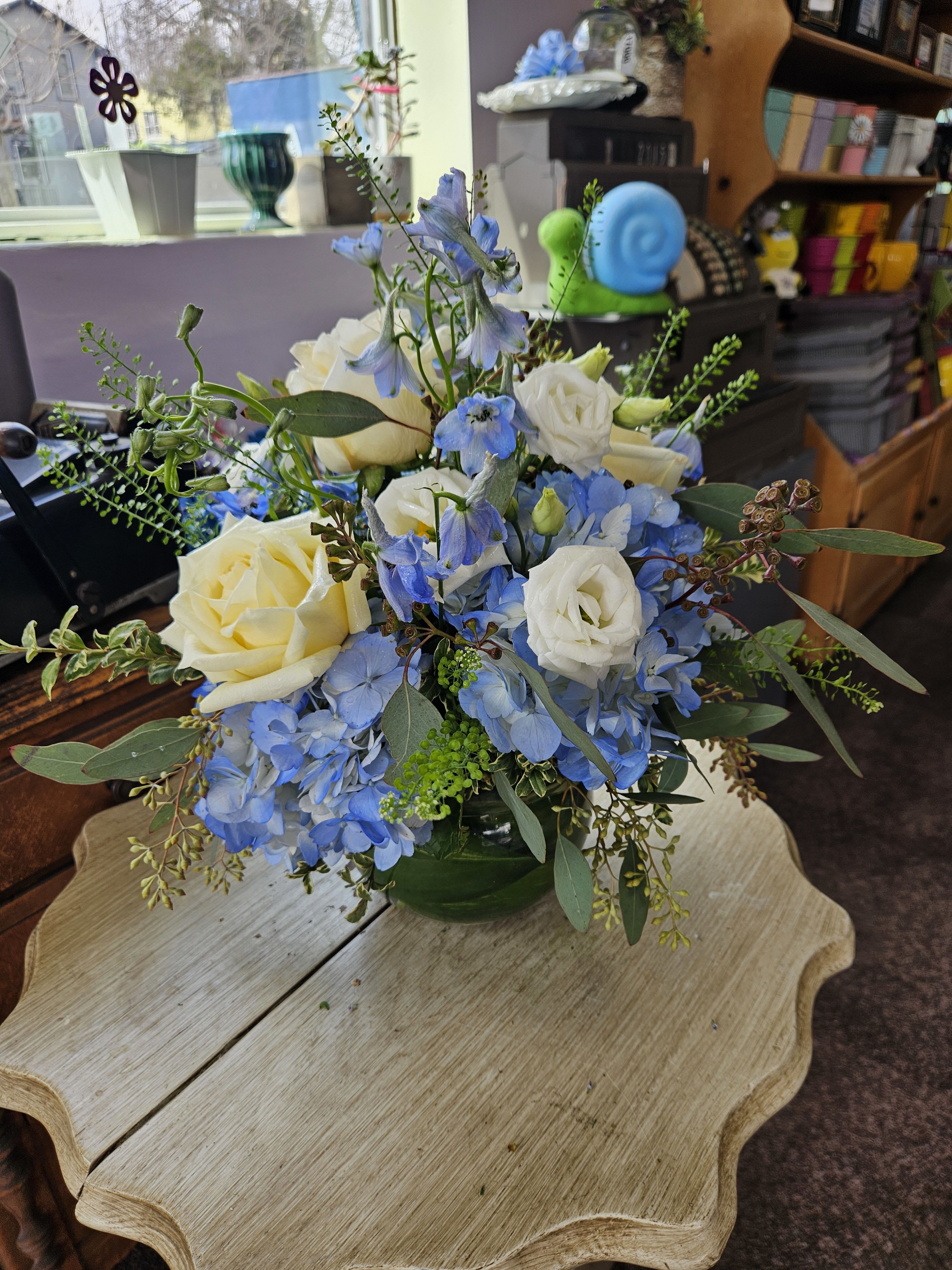 Low arrangement of white roses and blue flowers in a round green glass vase on a small table.