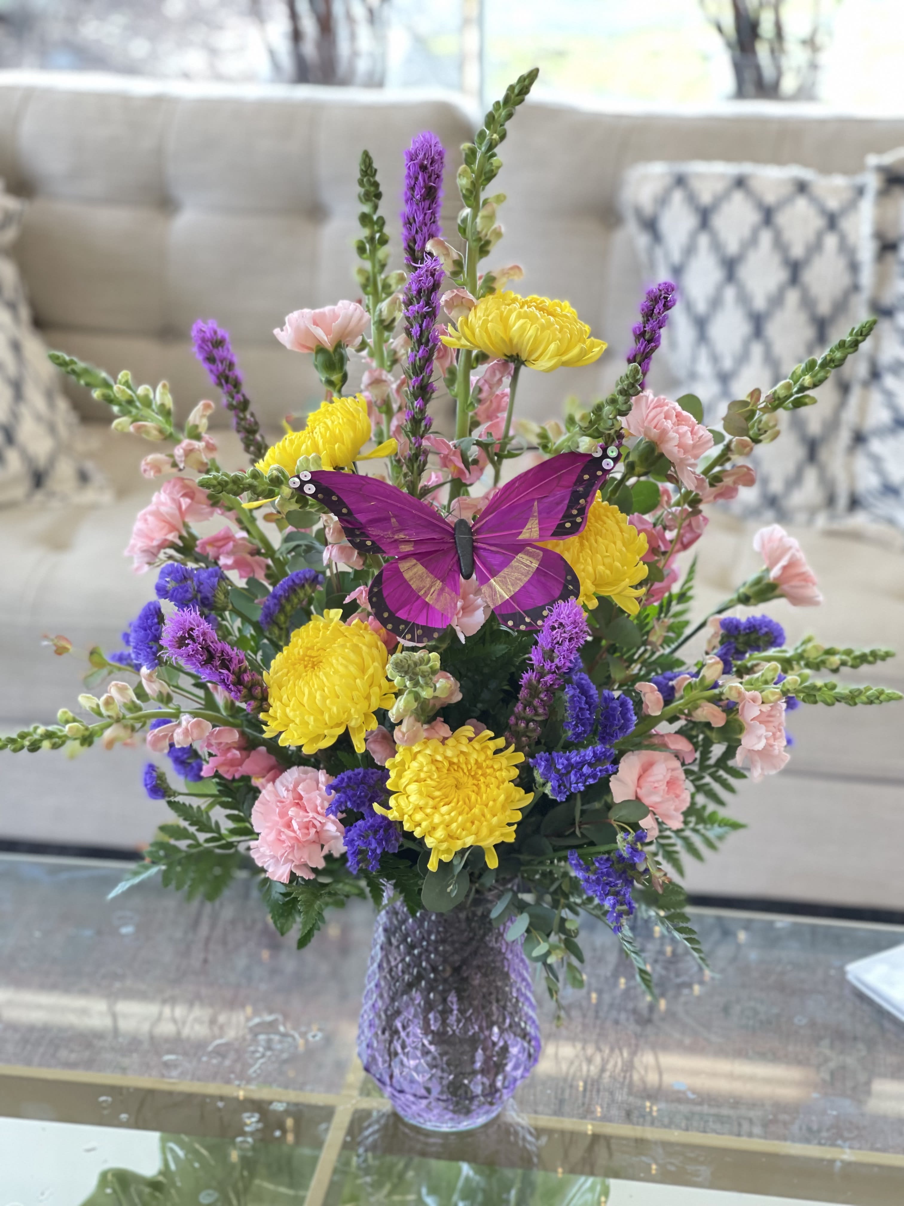 Bouquet of yellow, pink, and purple flowers in a purple vase with a butterfly decoration.