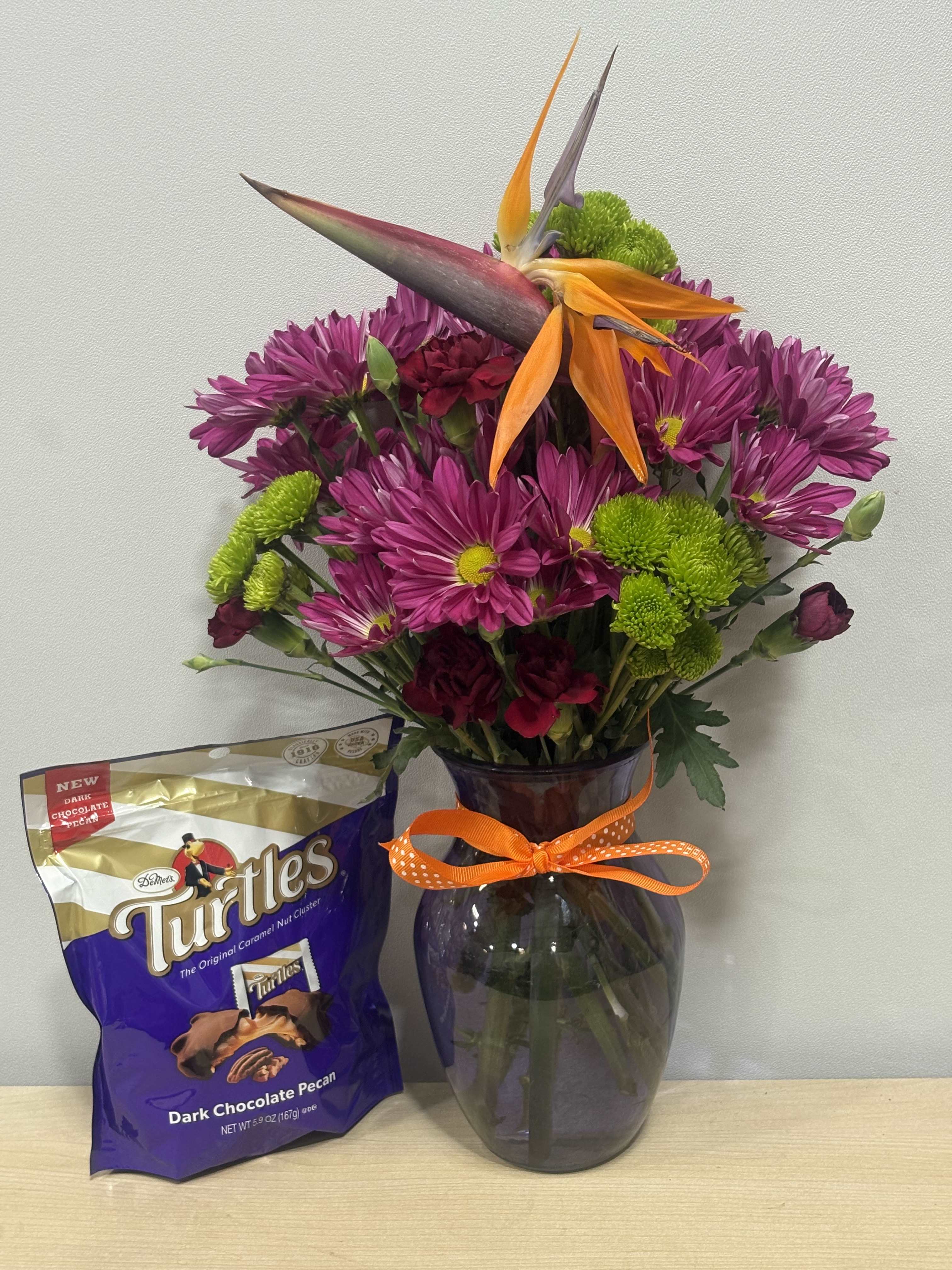 Purple and green flower arrangement in a glass vase with a chocolate candy bag