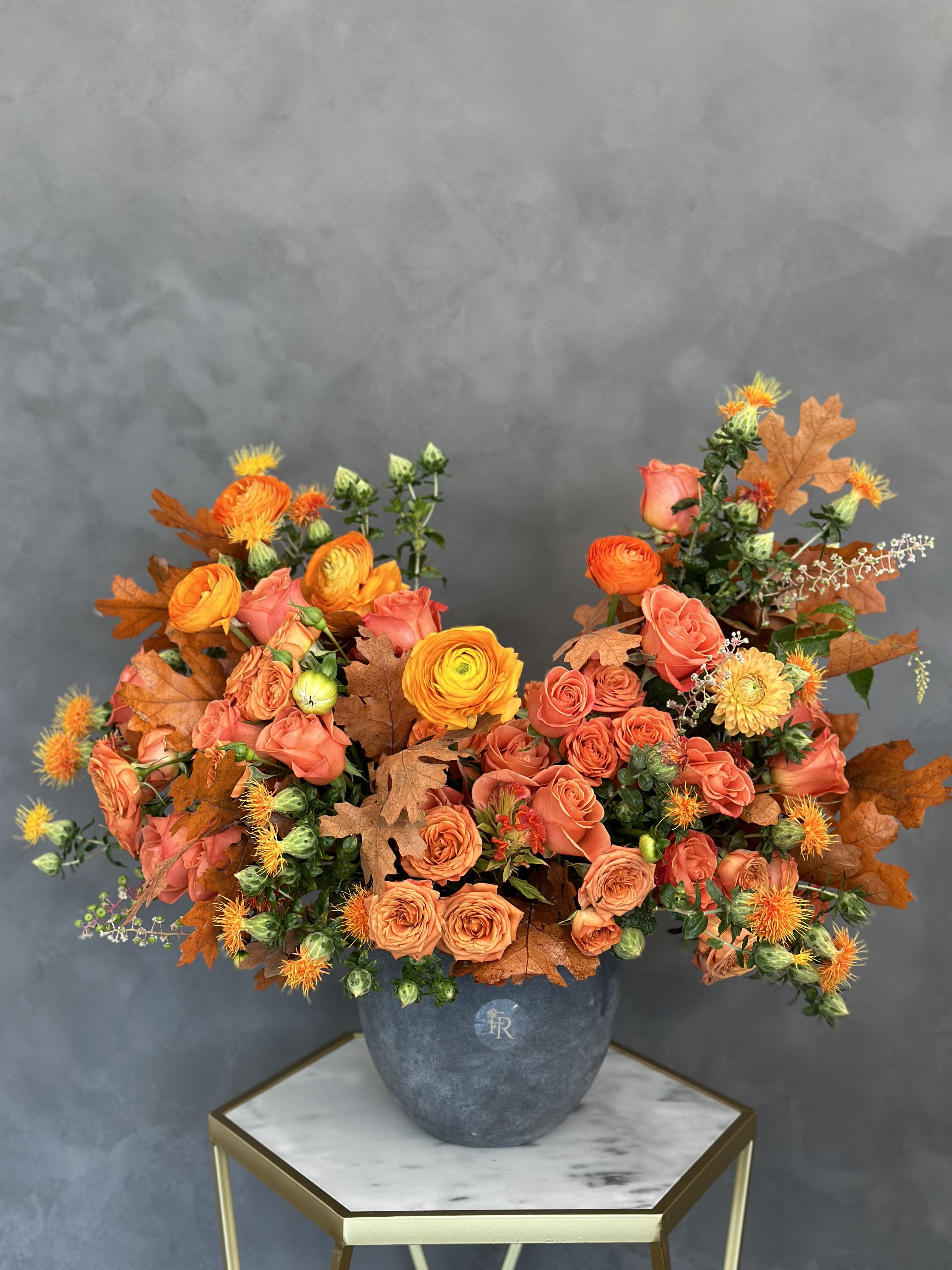 Orange and coral roses in a gray vase with autumn leaves