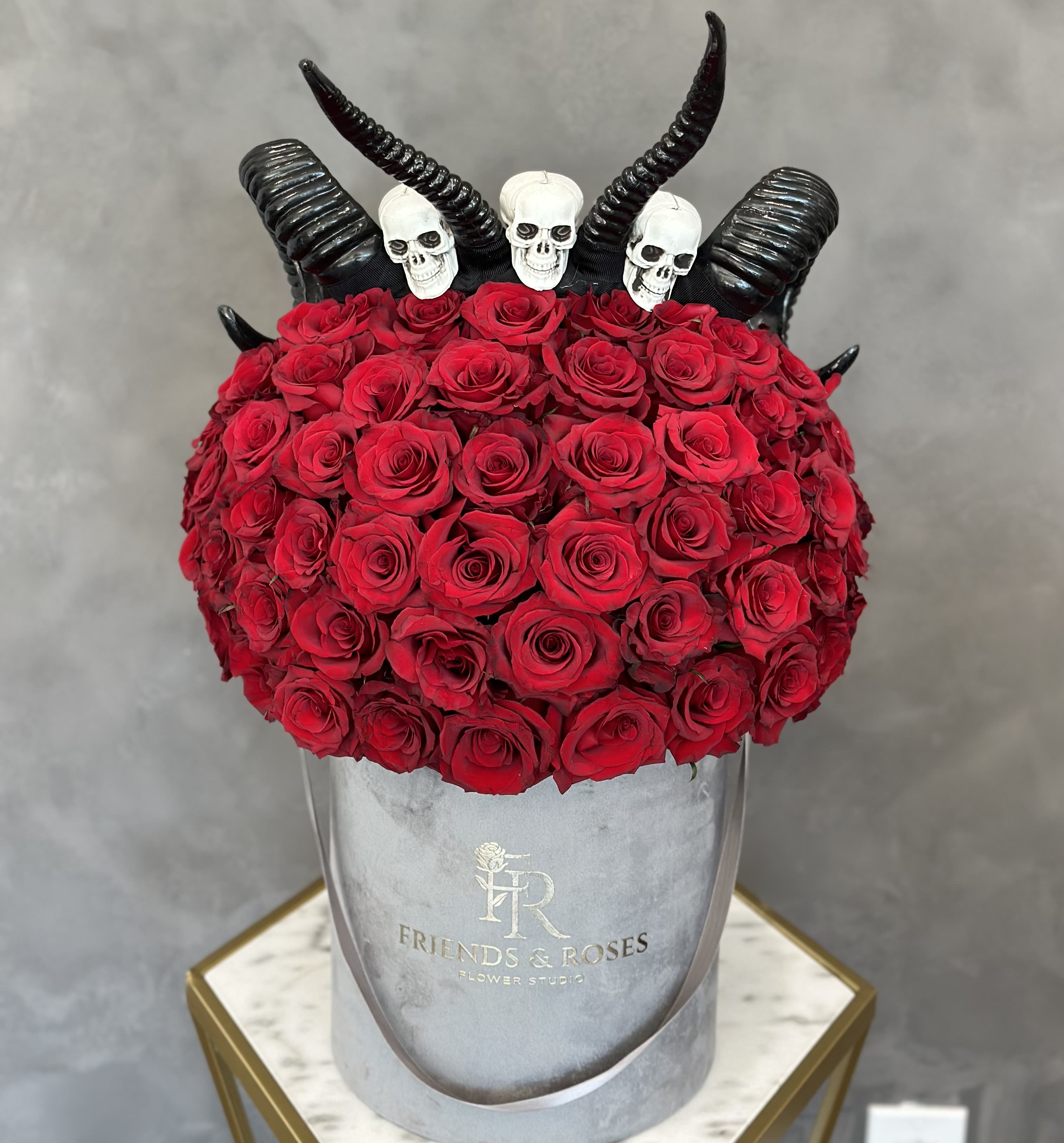 Red roses arranged in a gray hat box with black horn decorations and skull toppers