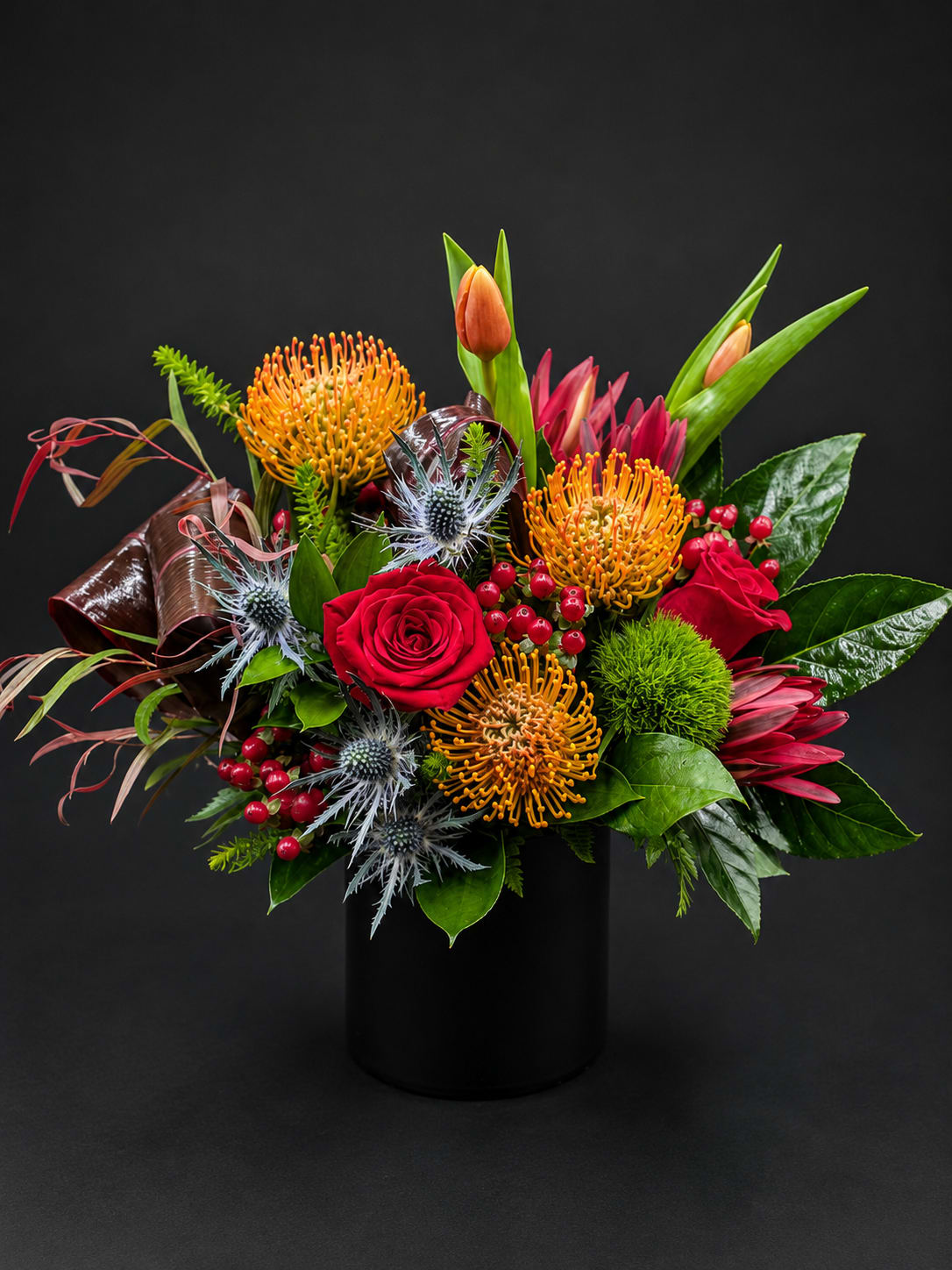 Tropical bouquet with red roses, orange pincushion blooms, and a black vase