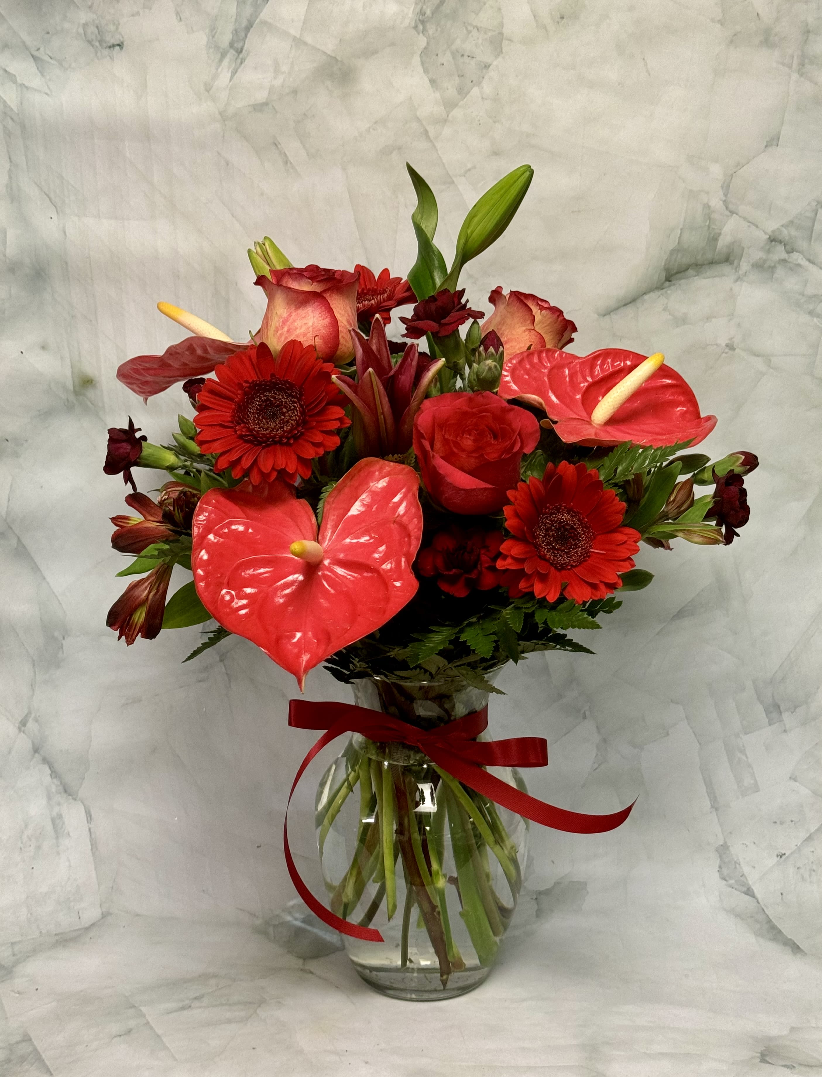 Red floral bouquet with anthuriums, roses, and gerbera daisies in a glass vase