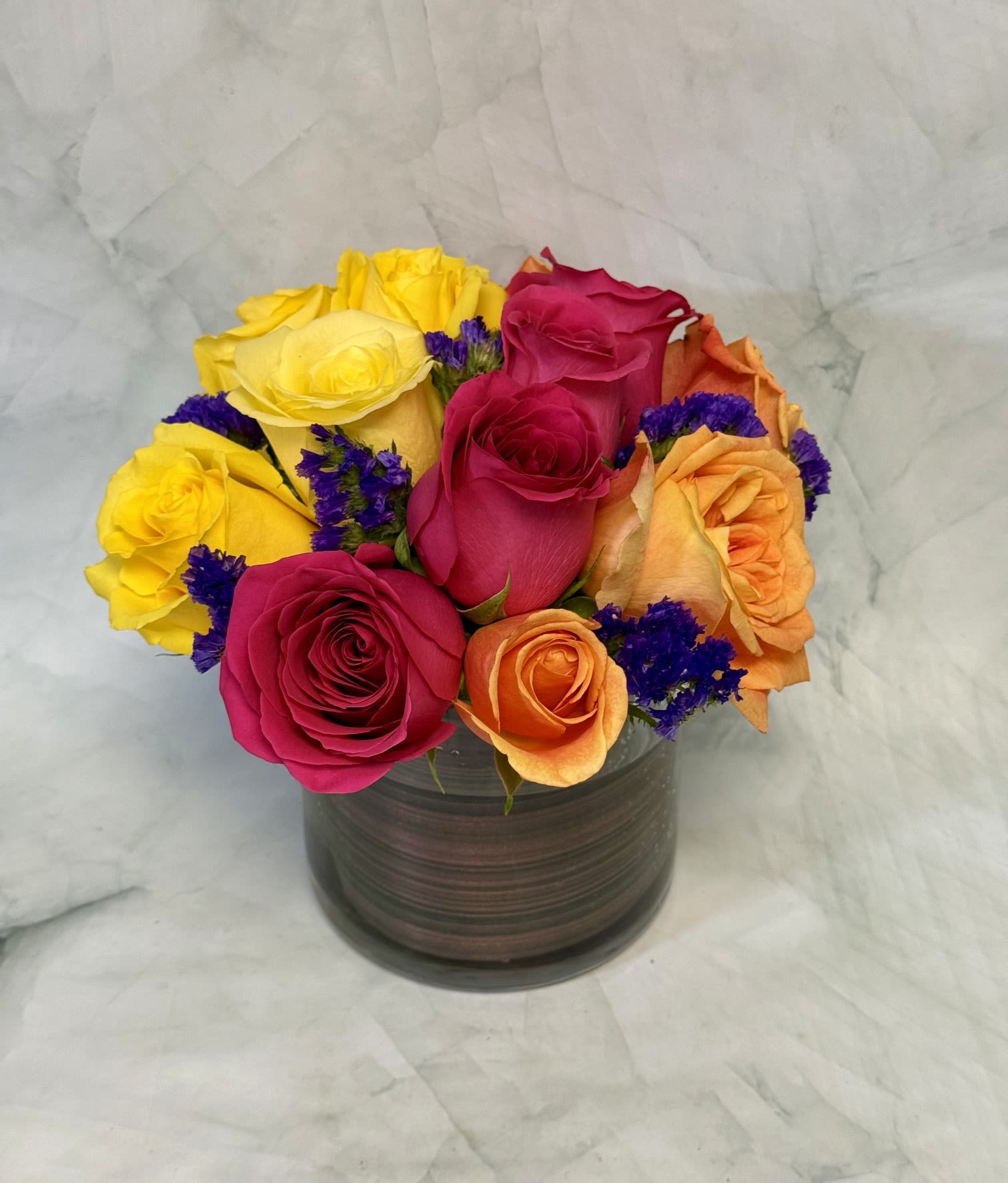 Modern Summer Roses - A dense, low cluster of summer roses in bright pink, peach-orange and sunny yellow-touched with pops of vivid purple-creates an immediate sense of celebration. Perfect as a summer bouquet or a graduation gift, this modern flower arrangement puts the roses front and center in a compact, rounded composition that reads joyful and festive for birthdays, congrats, or simply to make someone feel seen.  The stems are tightly packed into a clear square (or dark cylindrical) glass vase for a sleek, contemporary silhouette that highlights the saturated colors and lush rose heads. The arrangement is low and table-friendly, about the size of a dozen roses gathered into a cube-style container, with small purple filler flowers adding contrast and texture. This is the kind of cheerful, modern bouquet that makes the recipient feel genuinely celebrated-easy to place on a desk or dining table and designed for immediate visual impact.