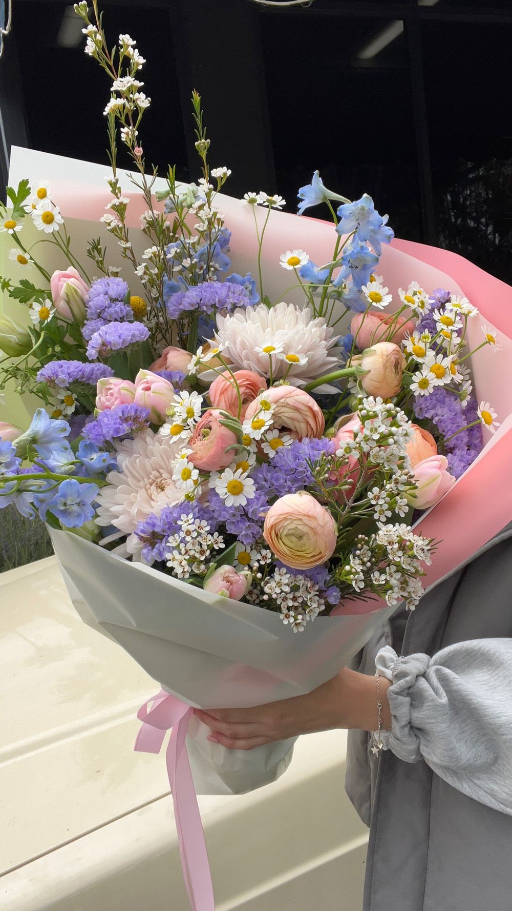 Pastel bouquet with pink ranunculus, white daisies, and blue flowers wrapped in pink paper