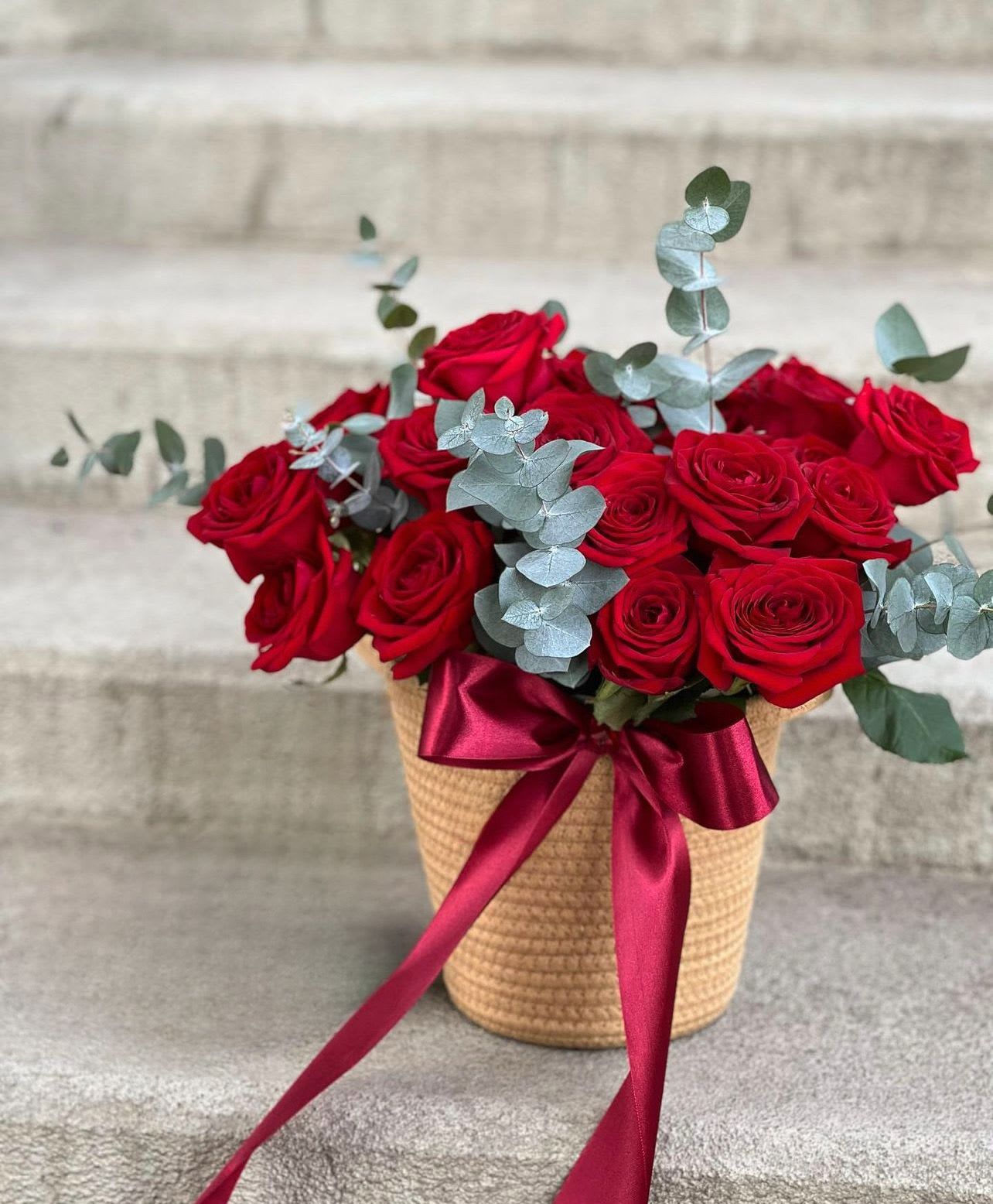 Red roses in a woven basket with a burgundy ribbon and eucalyptus