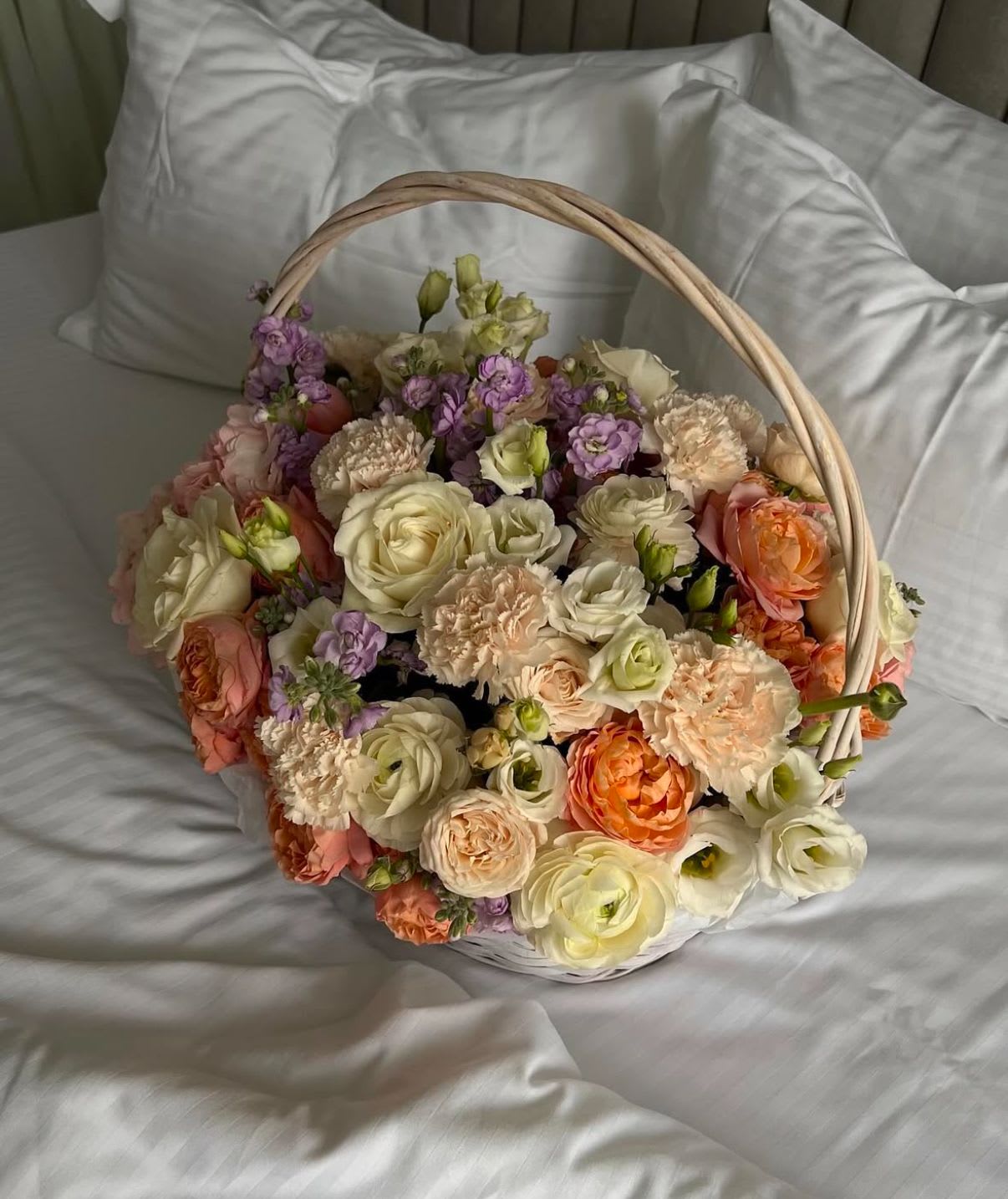 Basket of pastel roses, carnations, and purple blooms on white bedding