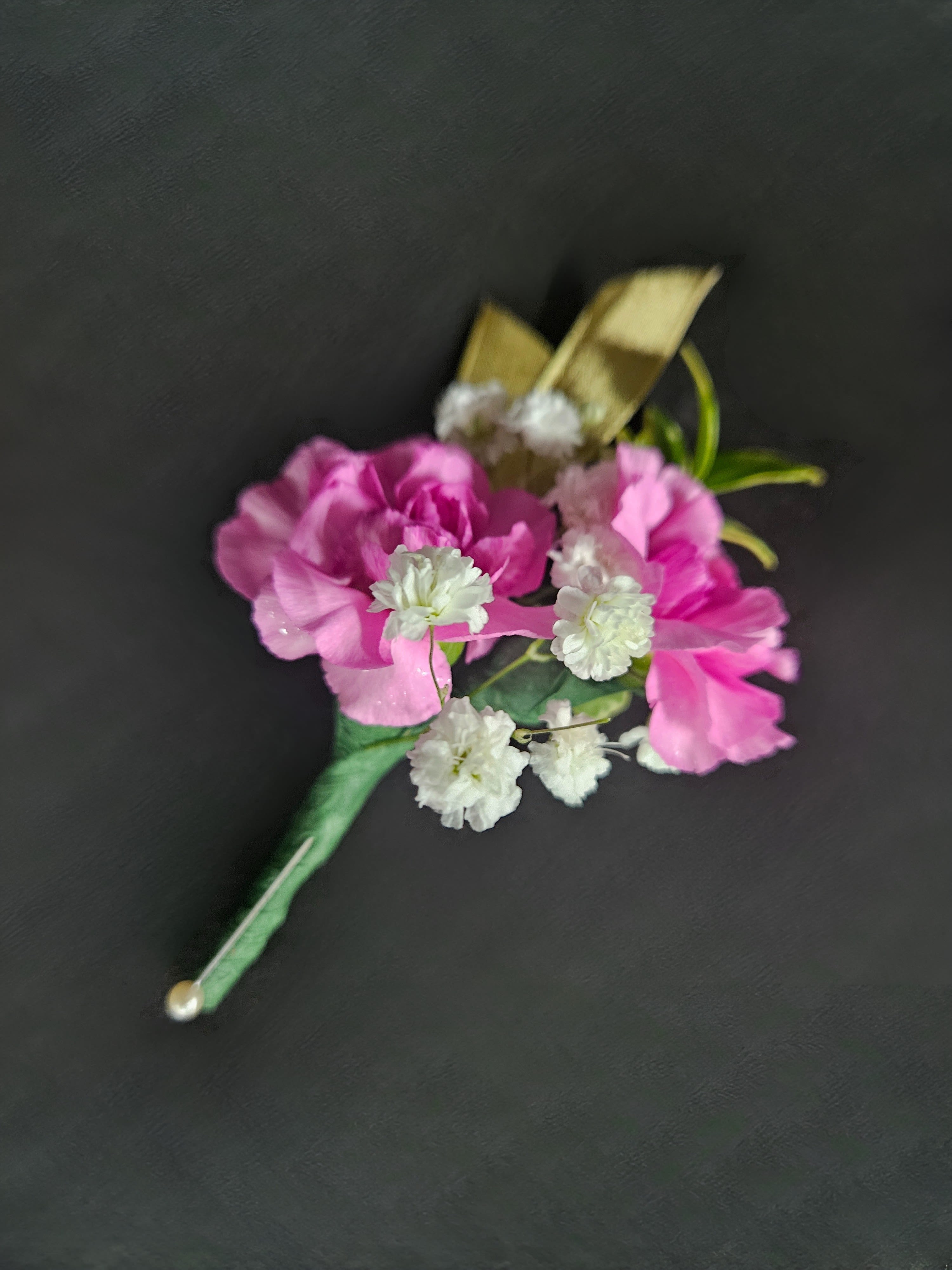 Pink carnation and white filler flower boutonniere with gold ribbon and pearl pin on a dark background