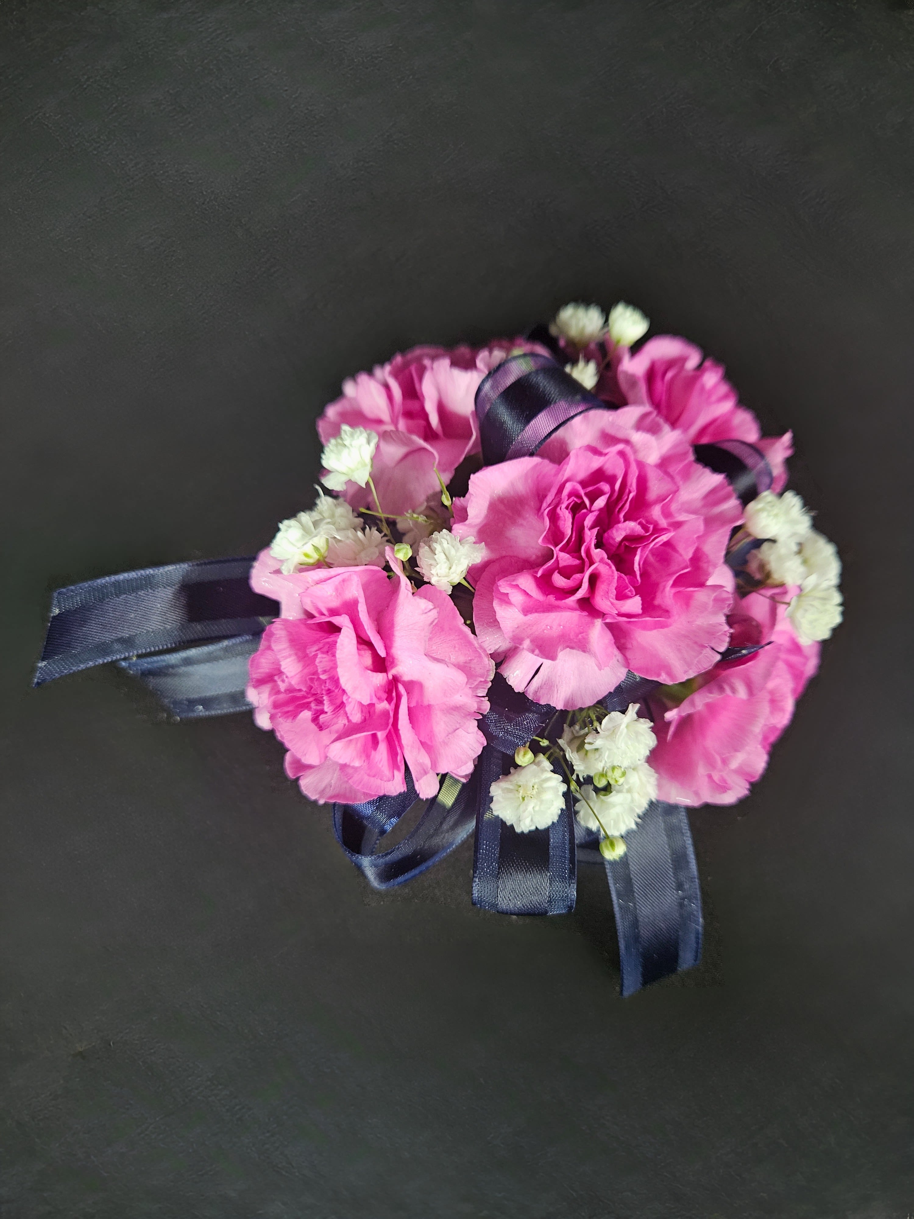Pink carnation corsage with white filler flowers and navy ribbon on a dark background