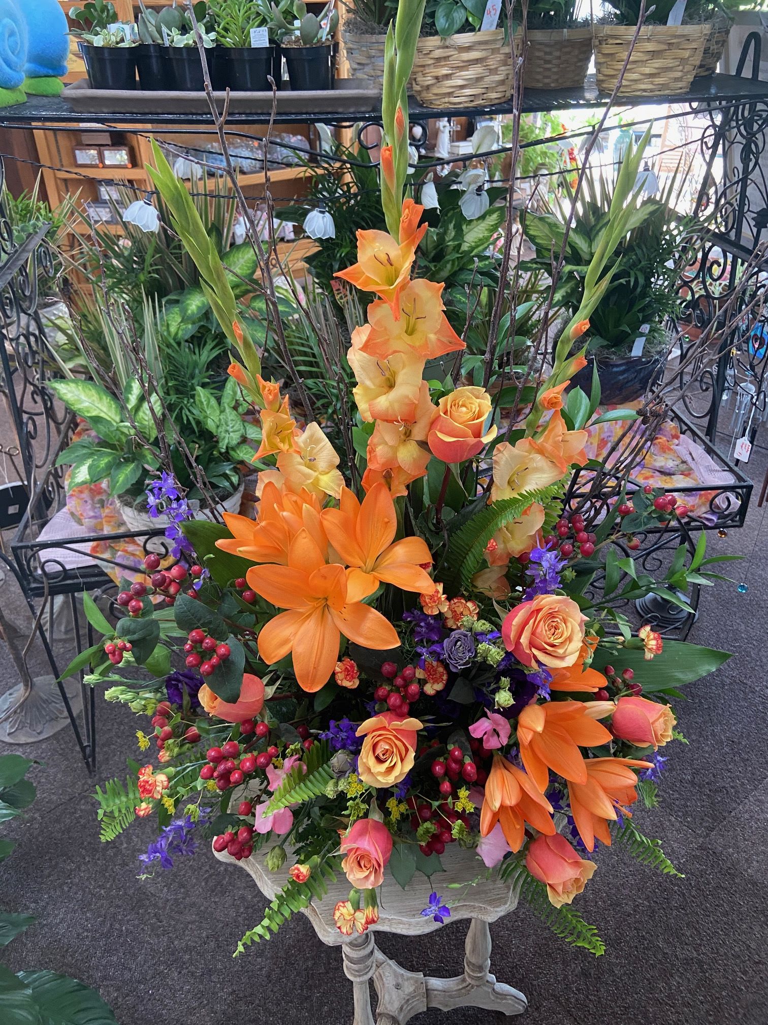 Tall arrangement of orange lilies and gladiolus with peach roses and purple flowers in a mixed display