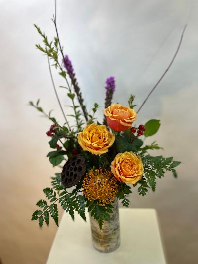 Orange roses in a tall glass vase with mixed greenery and purple accents