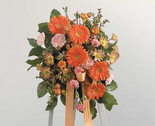 Tall standing spray of orange gerbera daisies and mixed flowers on an easel with peach ribbon.