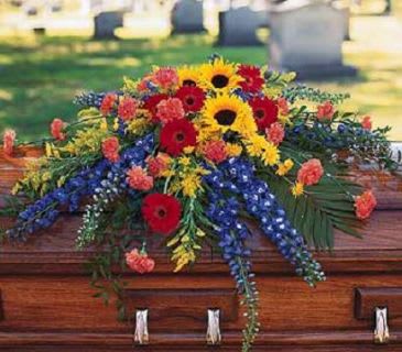 Bright mixed casket spray with sunflowers, red gerberas, and blue delphinium on a wooden casket