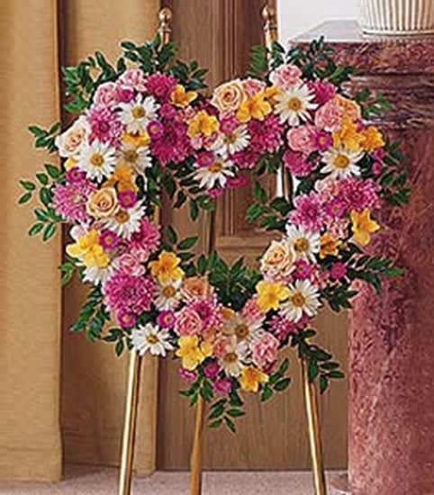 Heart-shaped standing wreath of pink, yellow, and white flowers on a gold easel.