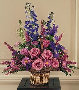 Tall basket arrangement of pink roses, purple irises, and lavender asters in a woven wicker basket