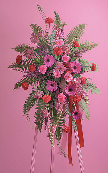 Tall pink and red standing spray with gerbera daisies, roses, carnations, and ferns on an easel.