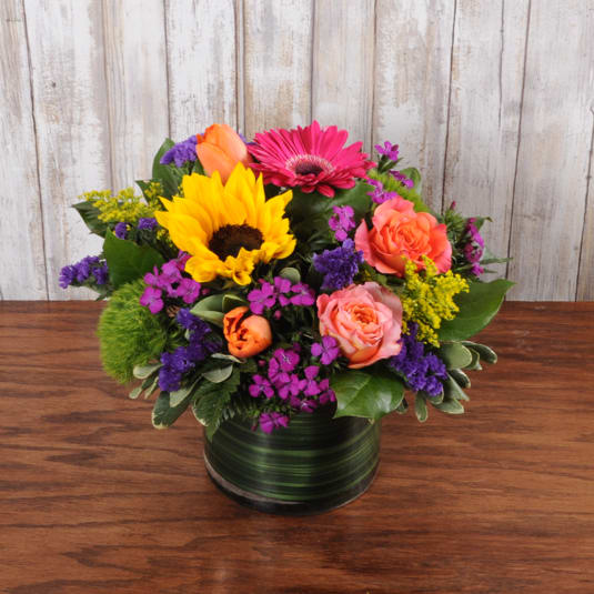 Low mixed bouquet with sunflower, pink gerbera, roses and purple flowers in a glass cylinder vase