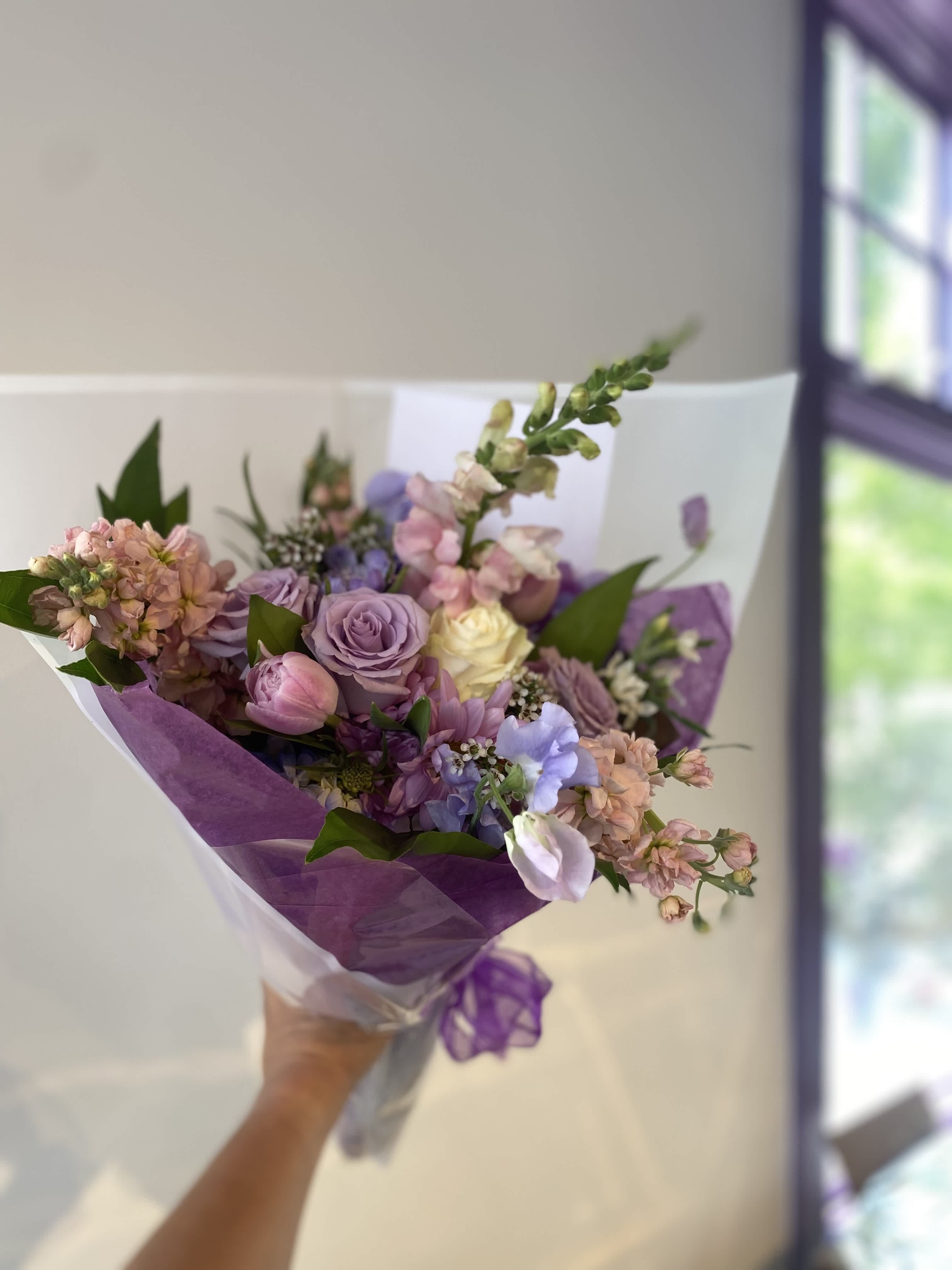 Handheld bouquet of lavender, pink, and cream flowers wrapped in purple paper