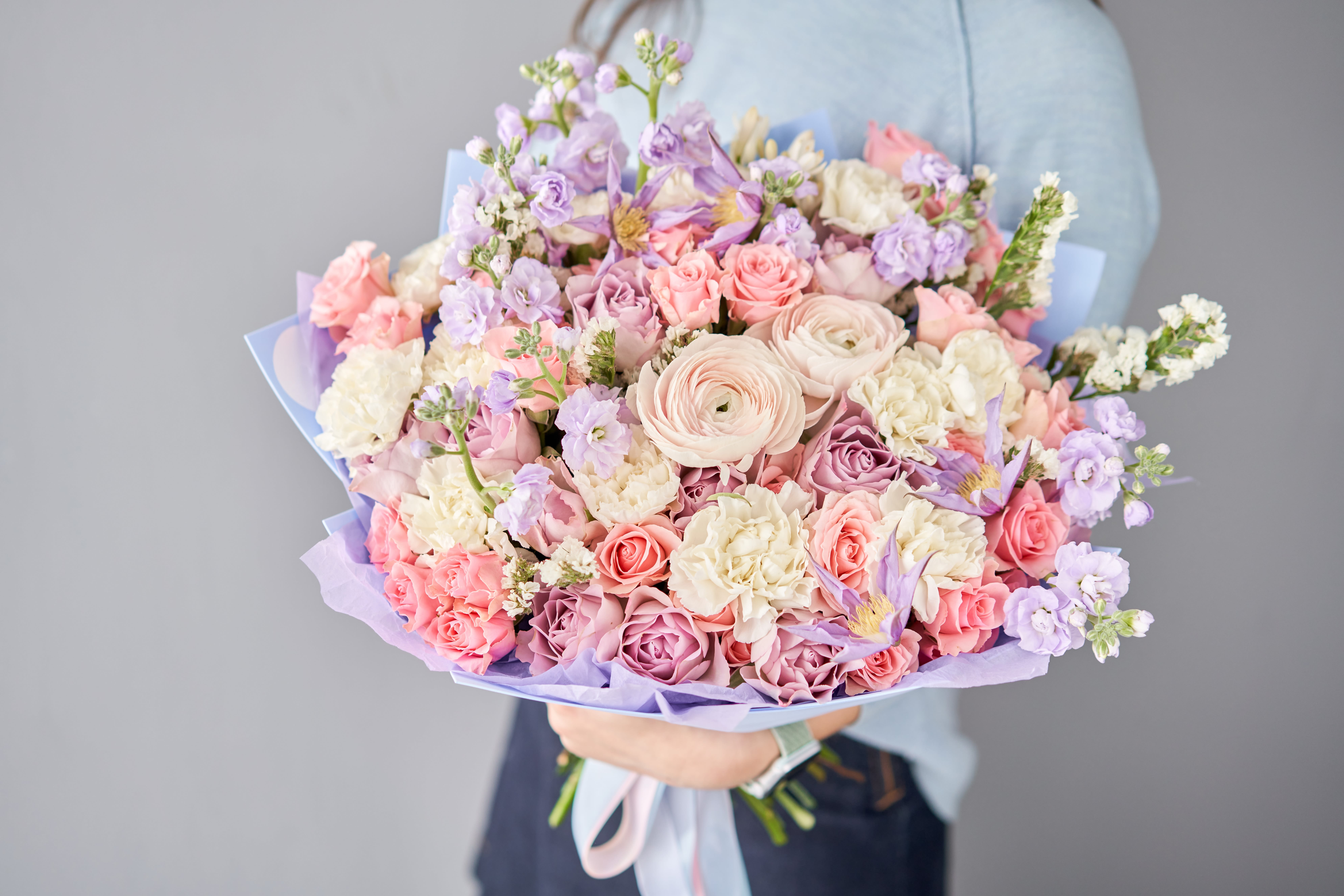 Large pastel bouquet of pink roses, white blooms, and lavender flowers wrapped in lilac paper, held in both hands