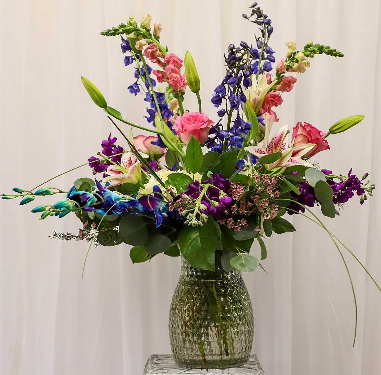 Tall mixed bouquet with lilies, roses, and purple blooms in a glass vase