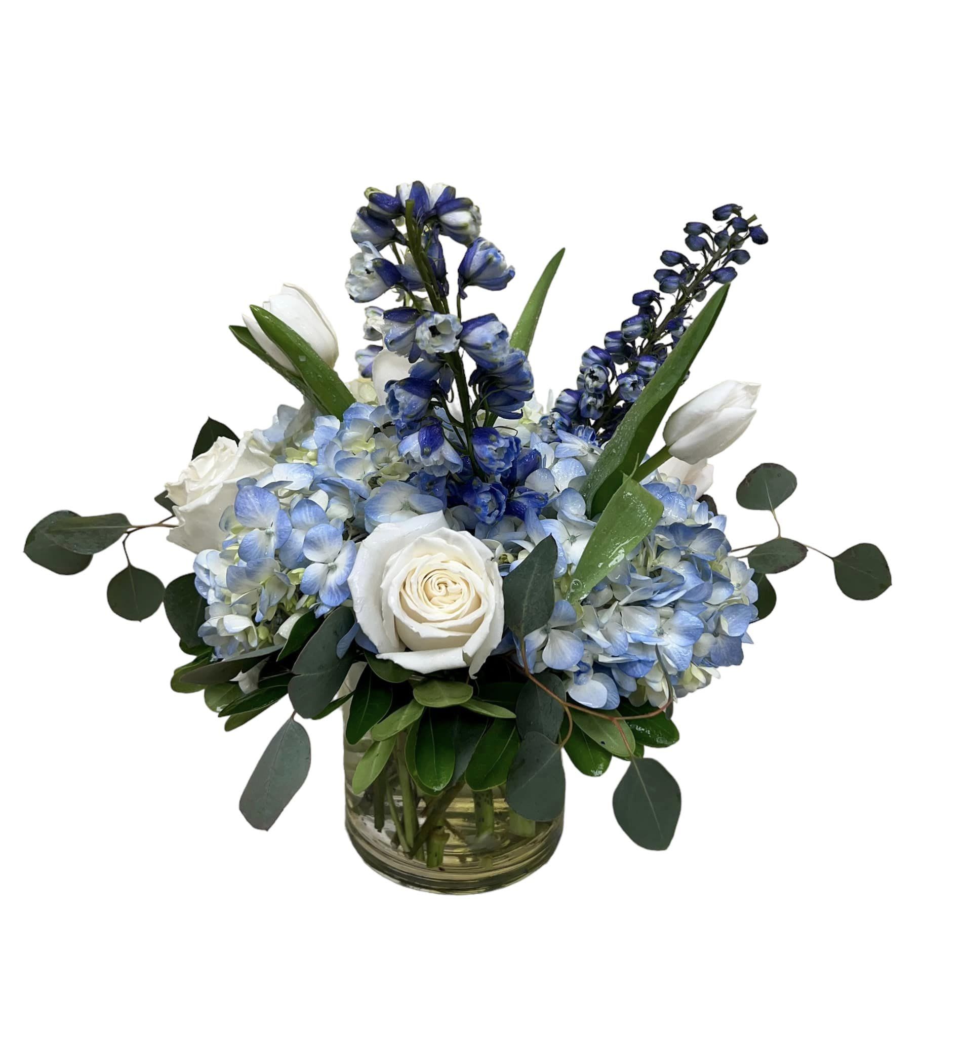 Blue hydrangeas, white roses, white tulips, and blue delphinium arranged in a clear glass cylinder vase