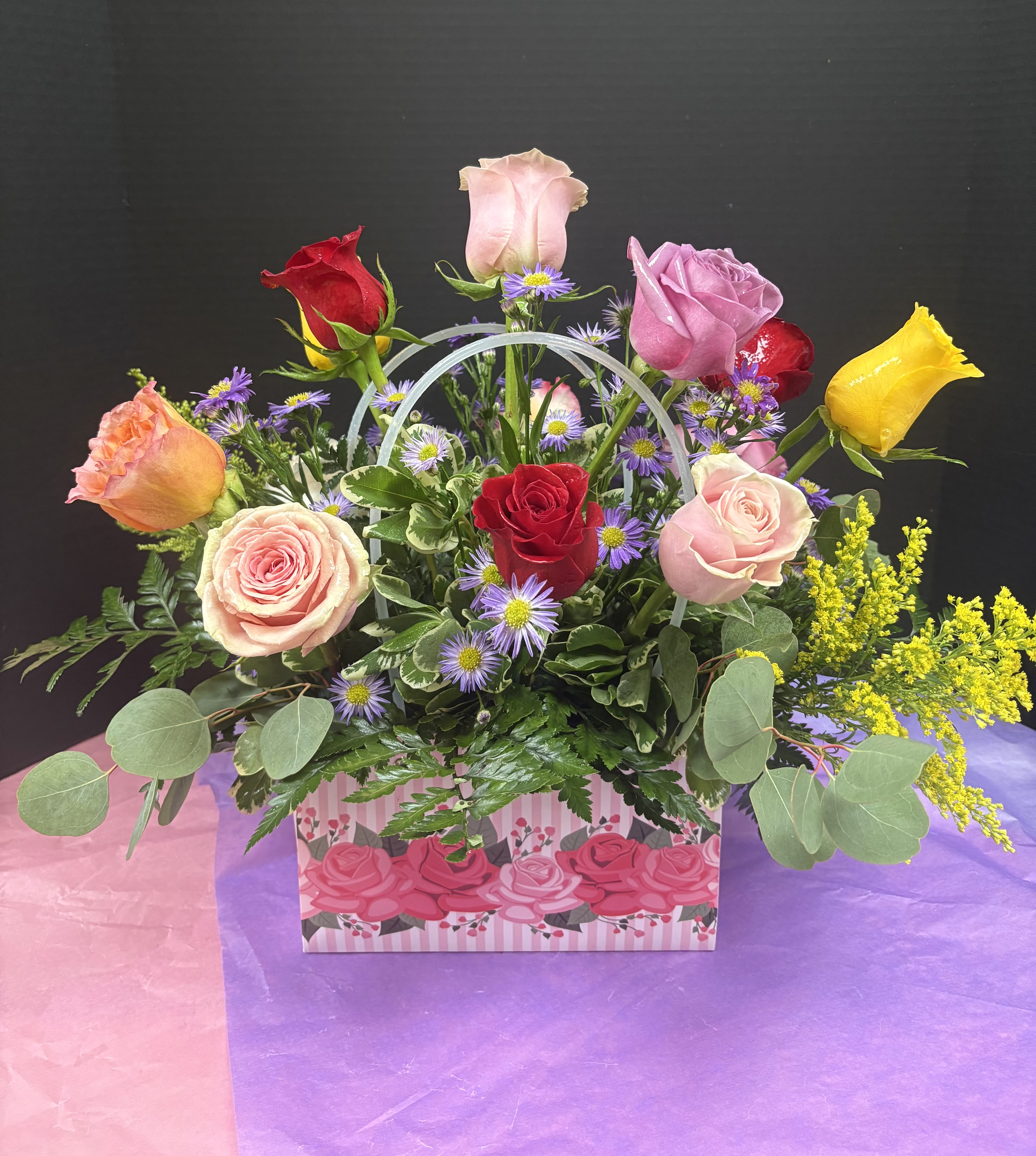 Rose Handbag  - A bright, giftable presentation of mixed-color roses that reads like a bouquet and a keepsake in one - perfect as a birthday bouquet, anniversary surprise, or just-because arrangement to say "I'm thinking of you." This flower arrangement for anniversaries and love and romance features clusters of pink, red, lavender and yellow roses paired with small purple aster-like blooms, arranged to create a lively, celebratory impression that also works for graduations, retirements, or to welcome a new baby.  Flowers are gathered densely inside a decorative pink-striped handbag gift box with rose graphics and a curved handle so it travels like a handbag and arrives ready to display. Textures include the soft, velvety rose petals, tiny daisy-like asters for contrast, touches of bright yellow filler and broad eucalyptus-like foliage that softens the composition. Size and configuration mirror a compact, hand-held presentation ideal for housewarming, thank-you, get-well, Mother's Day, Valentine's Day or Best Friends Day gifting.  This arrangement is made to delight the recipient the moment they see it - the kind of floral gift that feels thoughtful, celebratory, and unmistakably personal. Order for same-day delivery where available, and present the box as-is: no vase required, making it an easy, elegant choice for holiday visits, sympathy gestures, or a spontaneous "congrats" that brightens someone's day.