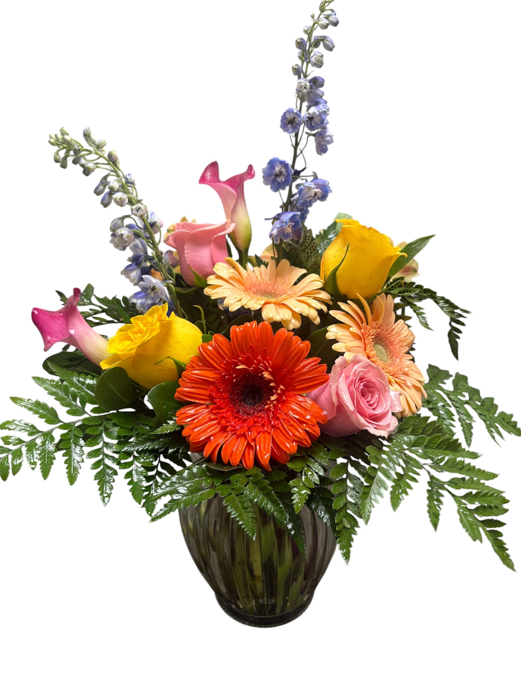 Neon Lights - Neon Lights delivers an unforgettable burst of color-perfect as a birthday bouquet, anniversary surprise, or a bright "just because" gift. This mixed-color flower arrangement features vivid orange gerbera daisies and peach gerberas paired with pink and yellow roses, pink calla lilies, and soaring blue delphinium spikes for dramatic height. The contrast between warm, sunlit tones and cool delphinium draws the eye and makes the recipient feel instantly cheered and celebrated.  Arranged in a glass vase with rounded, garden-style layering, the composition reads full and modern: lower blooms create a lush, textured base while the delphinium adds vertical elegance. Fern-like foliage fans gently around the base to soften the silhouette and give a freshly gathered-from-the-garden look. The scent is light and floral, and the vase gives this piece a polished finish appropriate for housewarming, graduation, retirement, or to say "I'm sorry" or "thank you."  Whether you're sending congratulations, welcoming a new baby, honoring Mother's Day, or offering sympathy with thoughtful color, Neon Lights suits many occasions across spring and summer and remains appropriate year-round. Vase dimensions and stems are built to present a generous, eye-catching display; for best results, change the water every two days and trim stems slightly when you refresh the vase to extend bloom life.
