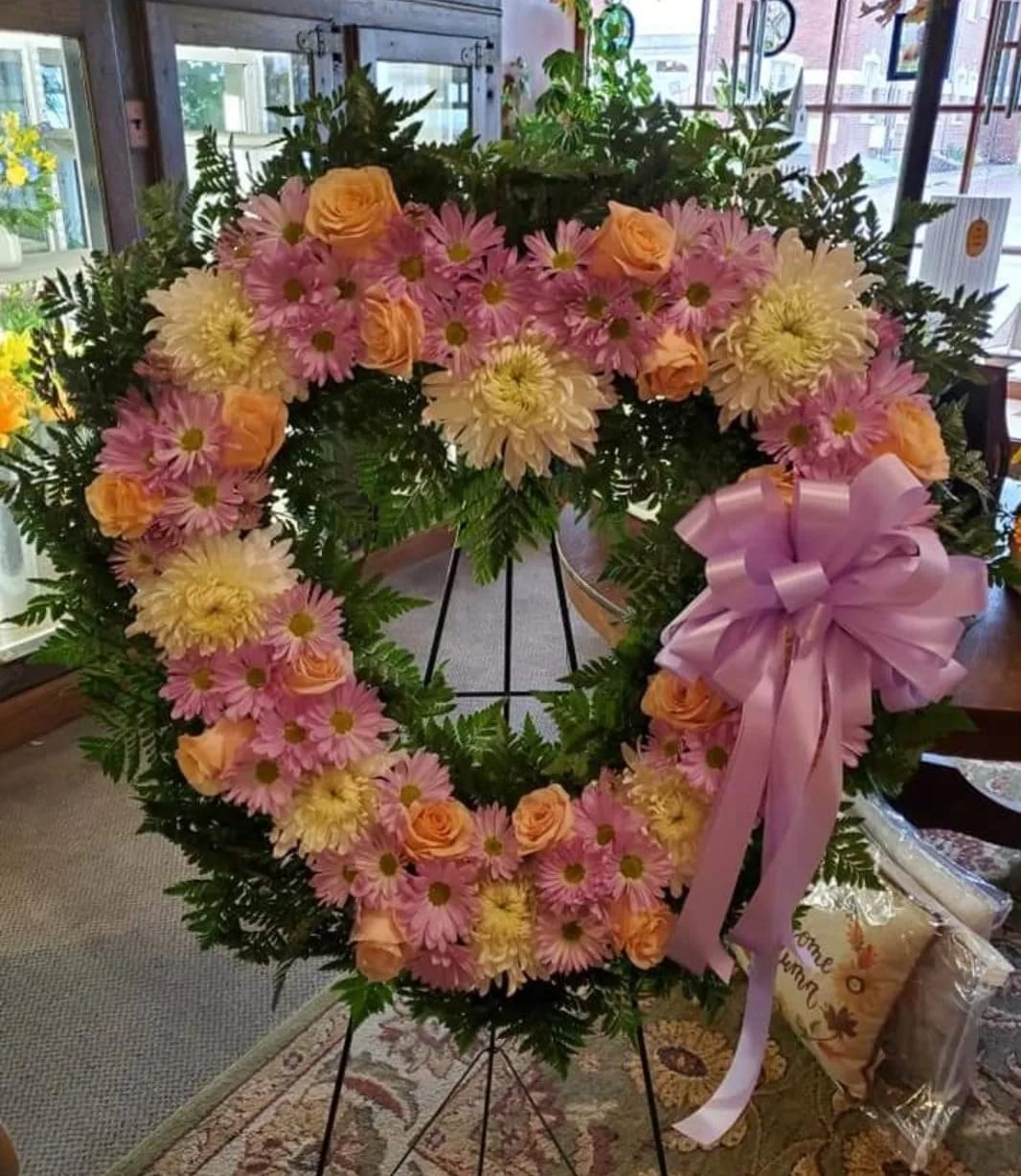 Forever in Our Heart Sympathy Standing Arrangement  - This splendid 24" heart-shaped wreath displays lovely shades of peach roses, lavender daisies, white magnums, greenery and a Lavender Bow. This gorgeous standing wreath is a beautiful way to express your deepest sympathies.