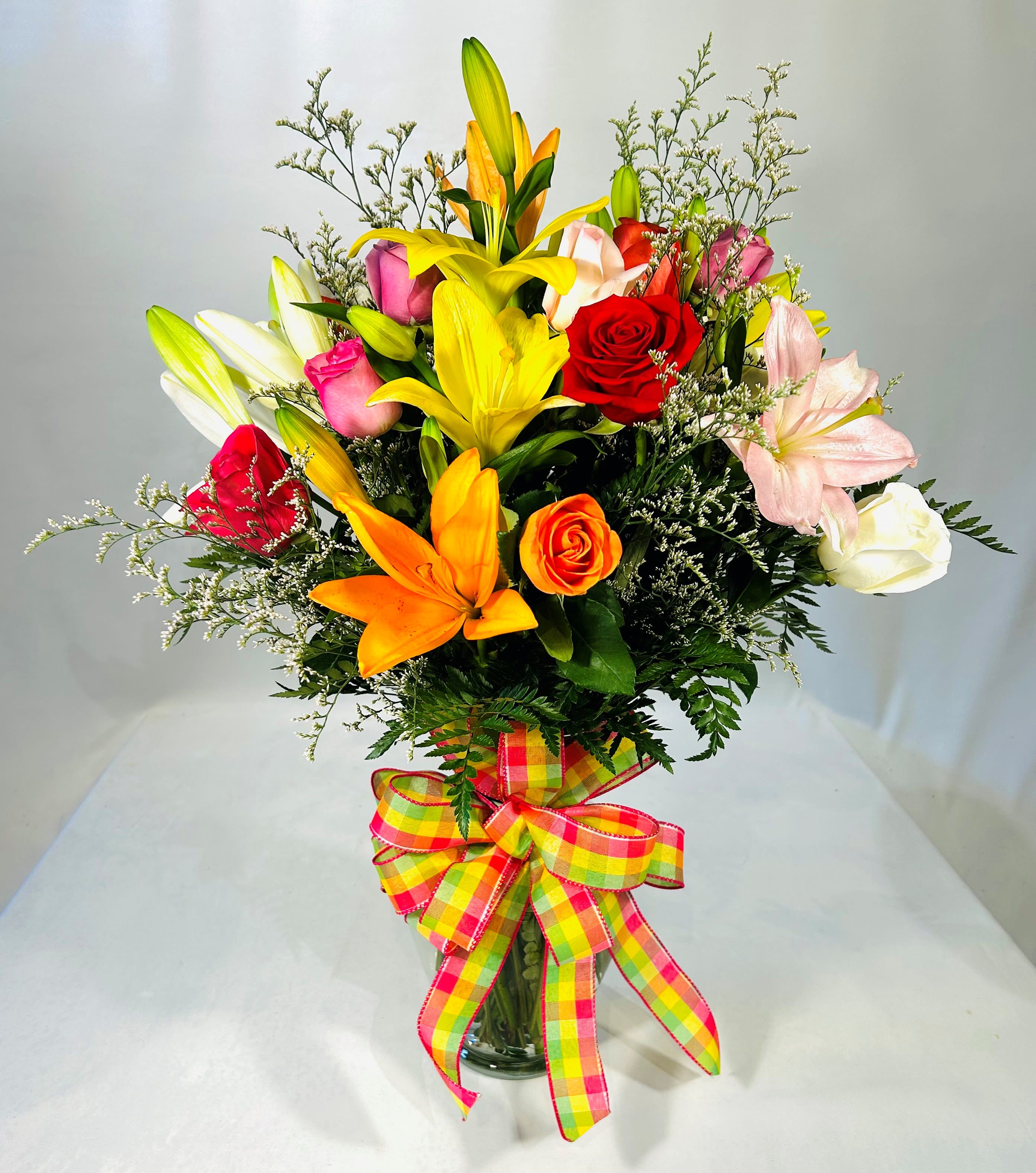 Summer Radiance - Sunlit yellow and orange lilies mingle with red, pink, orange and lavender roses to create a tall, joyful flower arrangement perfect for a summer birthday, Mother's Day surprise, or a bright "get well" or congratulations gift. The upright silhouette and lively mixed-color palette give the recipient an immediate lift-she'll feel noticed and celebrated the moment this bouquet arrives.  Large lilies and mixed roses are interspersed with delicate white filler blooms and lush greens for contrast and texture, densely clustered for a full, generous look. Stems are gathered in a classic clear glass vase and finished with a large multicolor plaid ribbon bow that adds a playful, celebratory touch. Sized to stand out on a table or bedside, this arrangement suits occasions from anniversaries, Father's Day and graduations to sympathy, "I'm sorry," or just-because moments-seasonal enough for summer and Mother's Day yet timeless for year-round gifting.