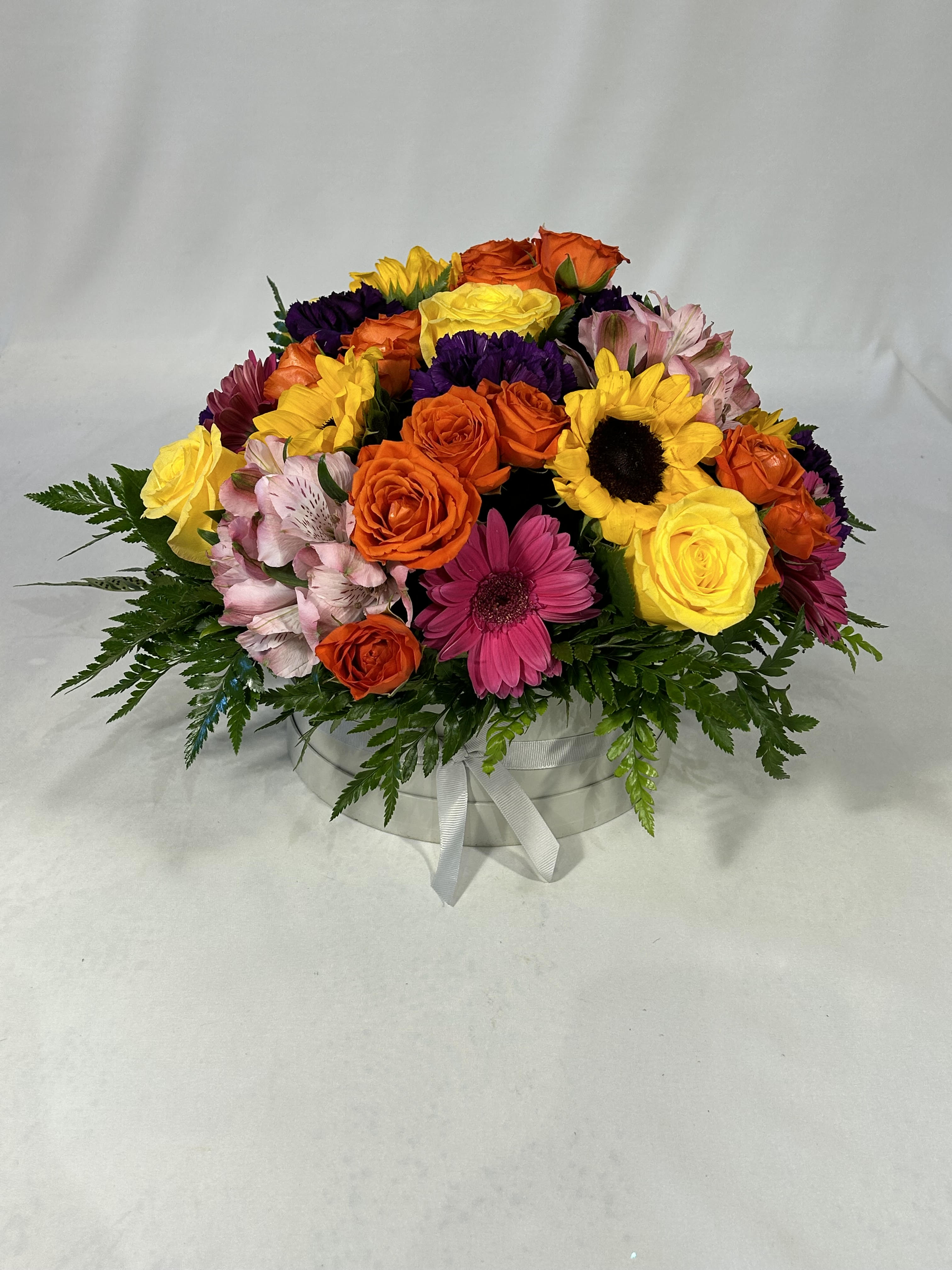Hatbox Delight - Bright, cheerful blooms spill from a round white hat box tied with a crisp white ribbon-an energetic flower arrangement perfect as a birthday bouquet, Mother's Day gift, or just-because surprise. The low, rounded design puts yellow sunflowers and orange roses front and center, while hot pink gerbera daisies and soft pink alstroemeria add playful warmth; deep purple carnations and chrysanthemums provide a striking contrast that makes the colors pop. This is the kind of arrangement that makes the person you're thinking of feel celebrated and seen, whether you're marking an anniversary, graduation, Father's Day, or sending get-well wishes.  Dense, tightly clustered flowers form a lush dome across the top of the white hatbox, with fern-like foliage framing the edges to give texture and a clean silhouette. Included blooms are alstroemeria, carnations, gerbera daisies, roses, spray roses, sunflowers, and chrysanthemums in a lively mix of yellow, orange, pink, purple, and mixed tones-an upbeat palette that also suits holidays, new baby congratulations, sympathy gestures, or a thoughtful thank-you. The compact design is a ready-to-display keepsake container that arrives full and abundant; order with same-day delivery when available to make any occasion feel a little brighter. 12" Tall