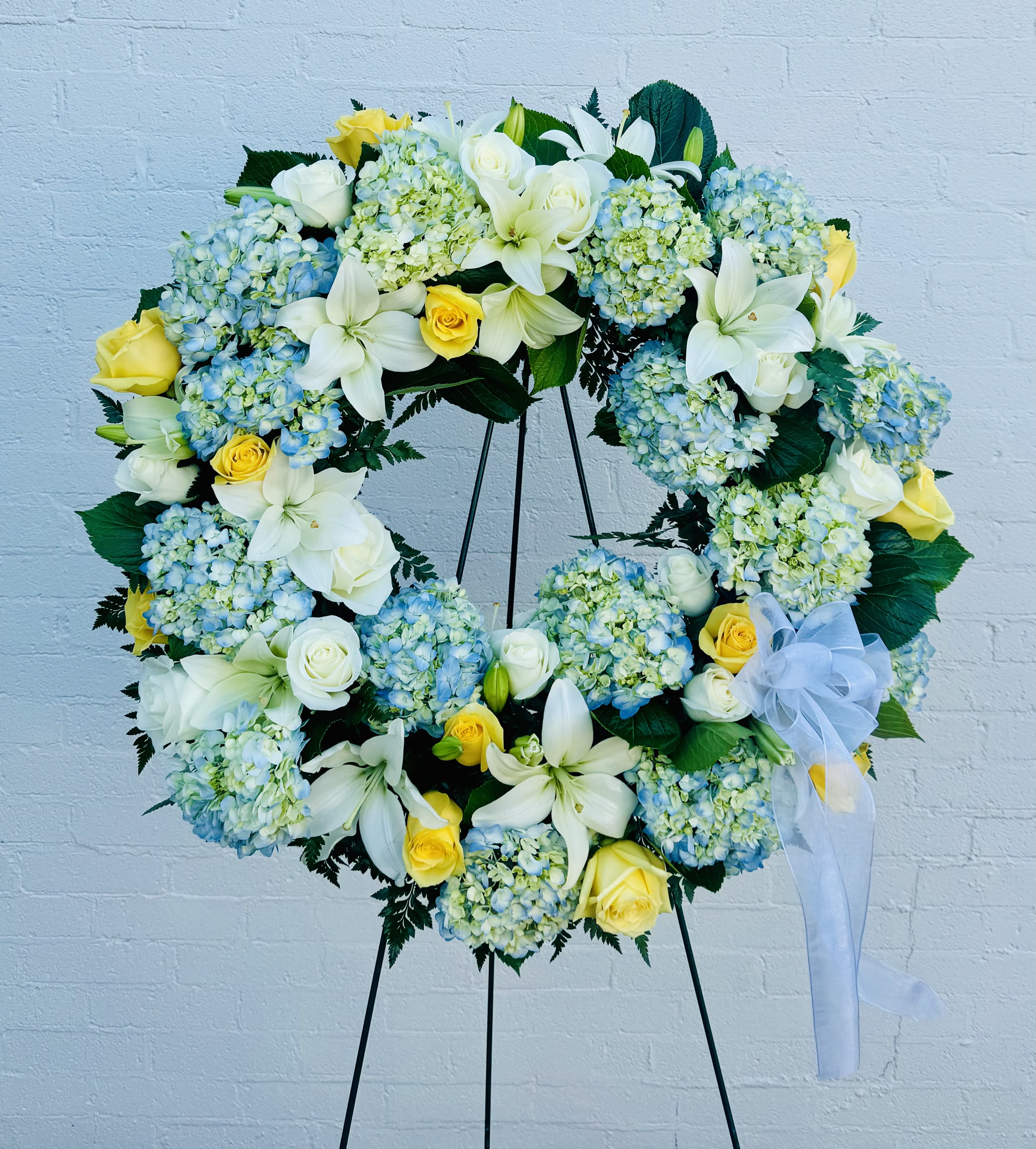 Large floral wreath with white lilies, yellow roses, and blue hydrangeas on a stand