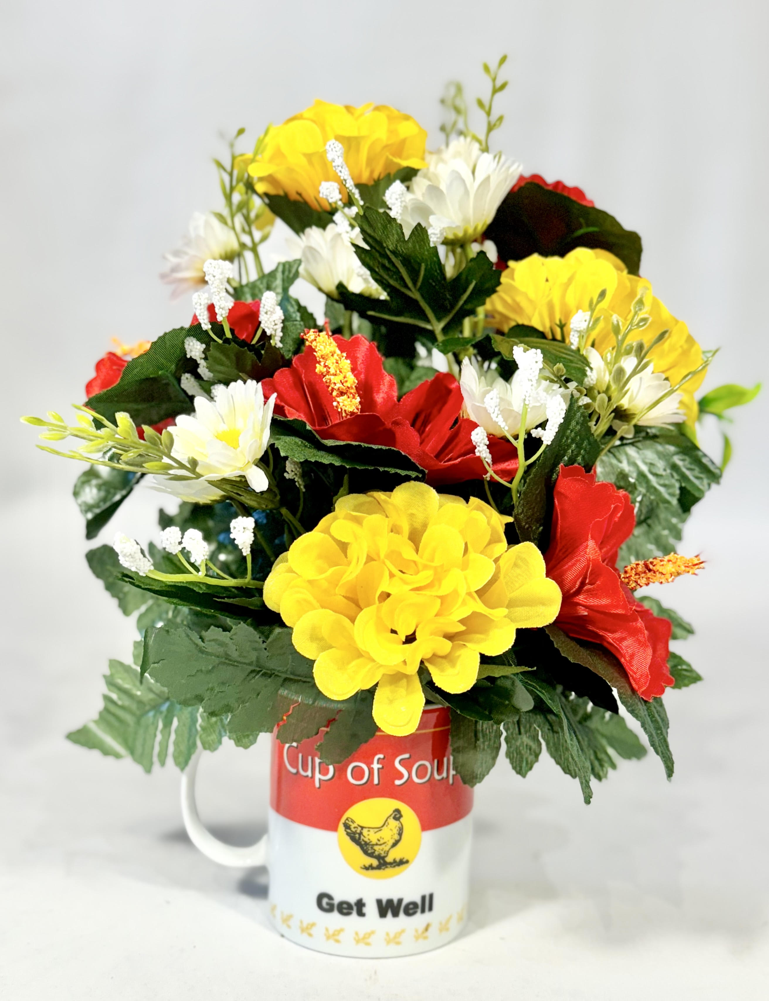 Get Well "Cup of Soup" Silk Arrangement - Get Well "Cup of Soup" silk arrangement is a bright and cheery display to help someone feel better. This mug can be sent as a Get Well Soon floral arrangement and then can be used afterwards as a soup mug or coffee mug.  10" H
