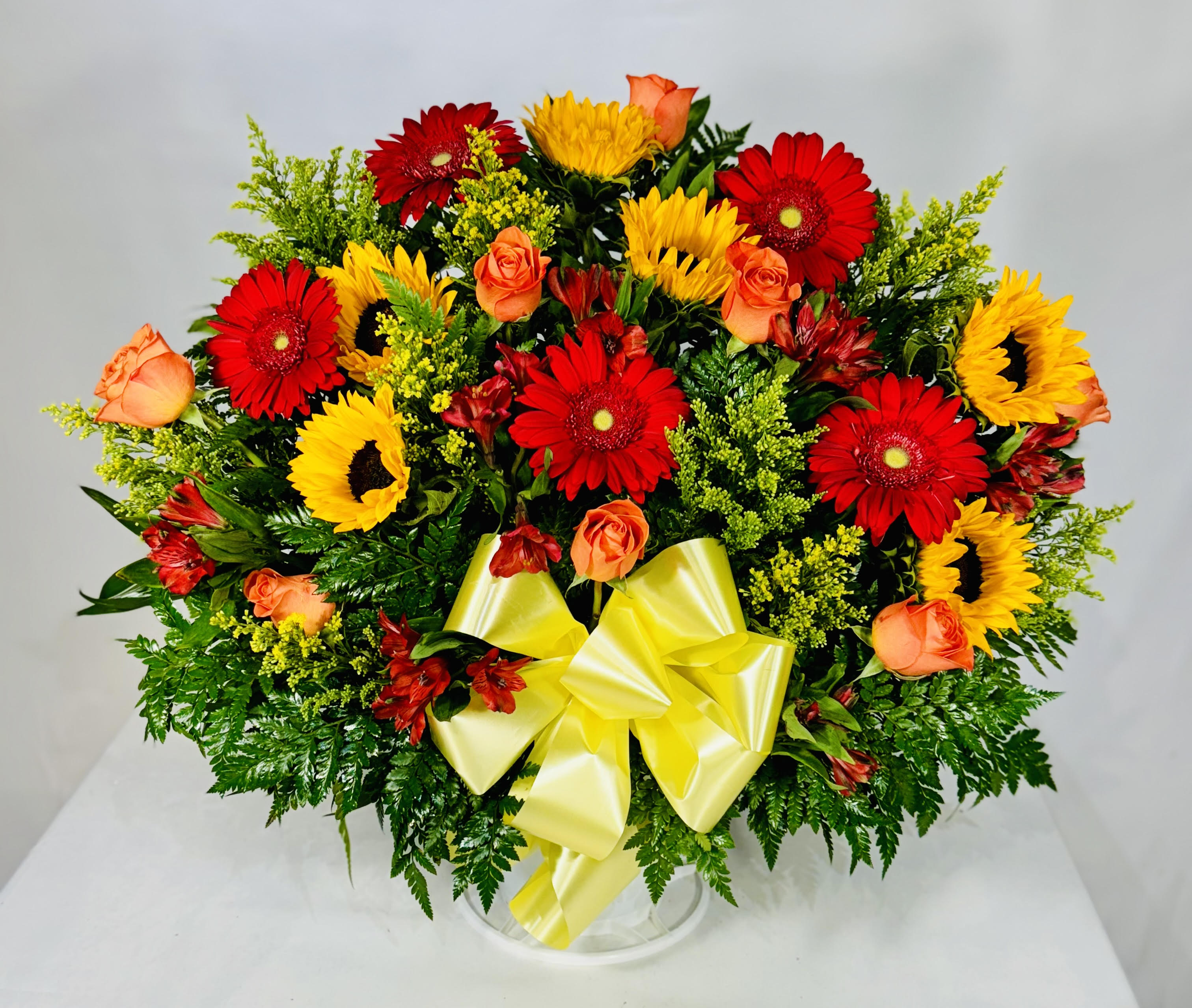 Mixed bouquet of red gerbera daisies, yellow sunflowers, and orange roses in a clear vase