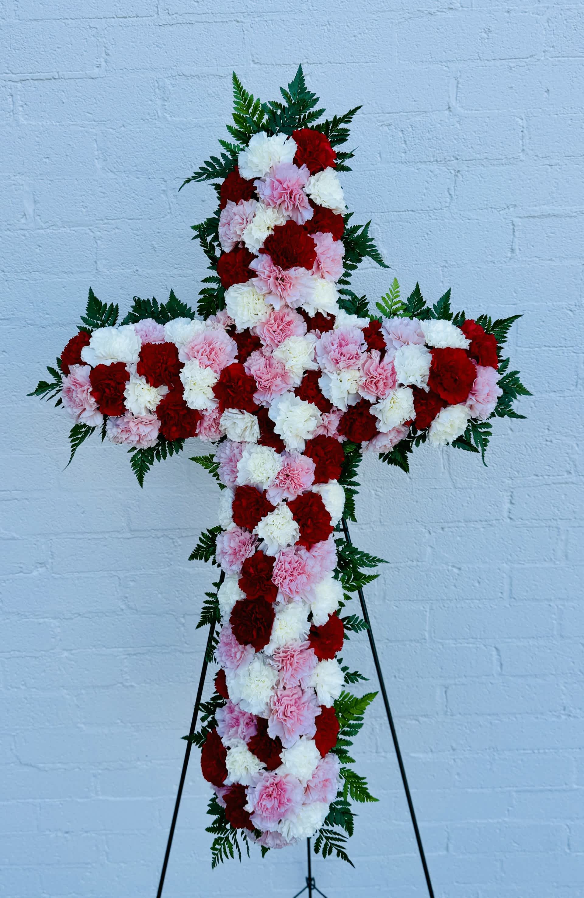  Red, White and Pink Carnation Standing Cross  - A densely covered standing cross of red, pink and white carnations that offers a reverent, devotional focal point for sympathy and funeral services. The close-set blooms create a solid, bold cross silhouette-deep red carnations add depth while soft pinks and whites bring a comforting, gentle presence-so the person you're honoring will be remembered with dignity and warmth.  Fern fronds peek just beyond the edges to soften the outline and add a touch of green texture, and the arrangement is mounted on a sturdy black metal easel for formal display at services or memorials. Finished size is approximately 22" wide by 34" high (dimensions do not include easel height), making it a meaningful tribute that reads clearly from the front pew and photographs beautifully for keepsakes. Colors of Carnations can be changed, please let us know in notes section on ordering or call and order and let us know. 