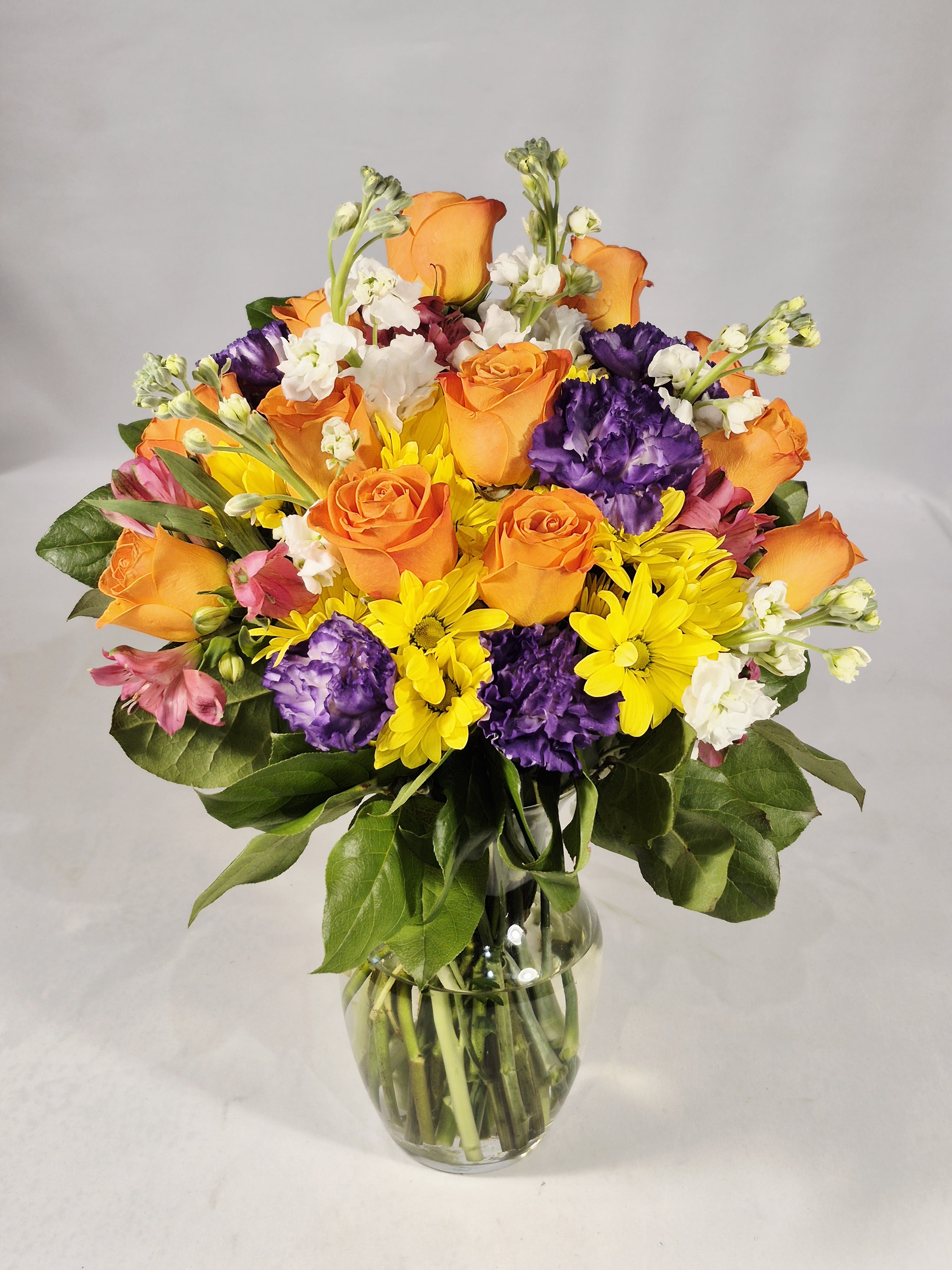Mixed bouquet of orange roses, purple carnations, and yellow daisies in a glass vase