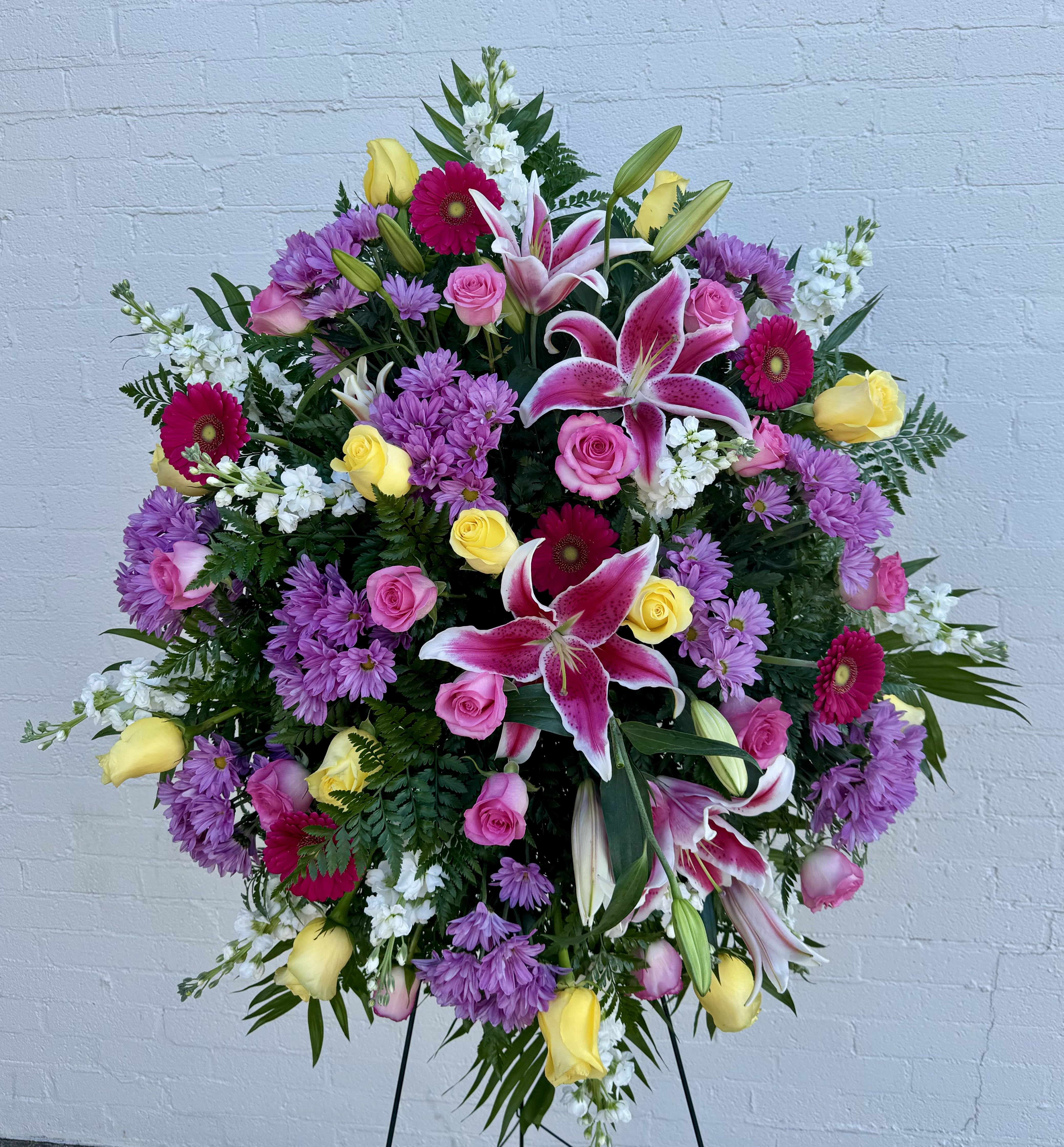 Large standing spray with pink lilies, roses, yellow roses, purple mums, and white accents on an easel.