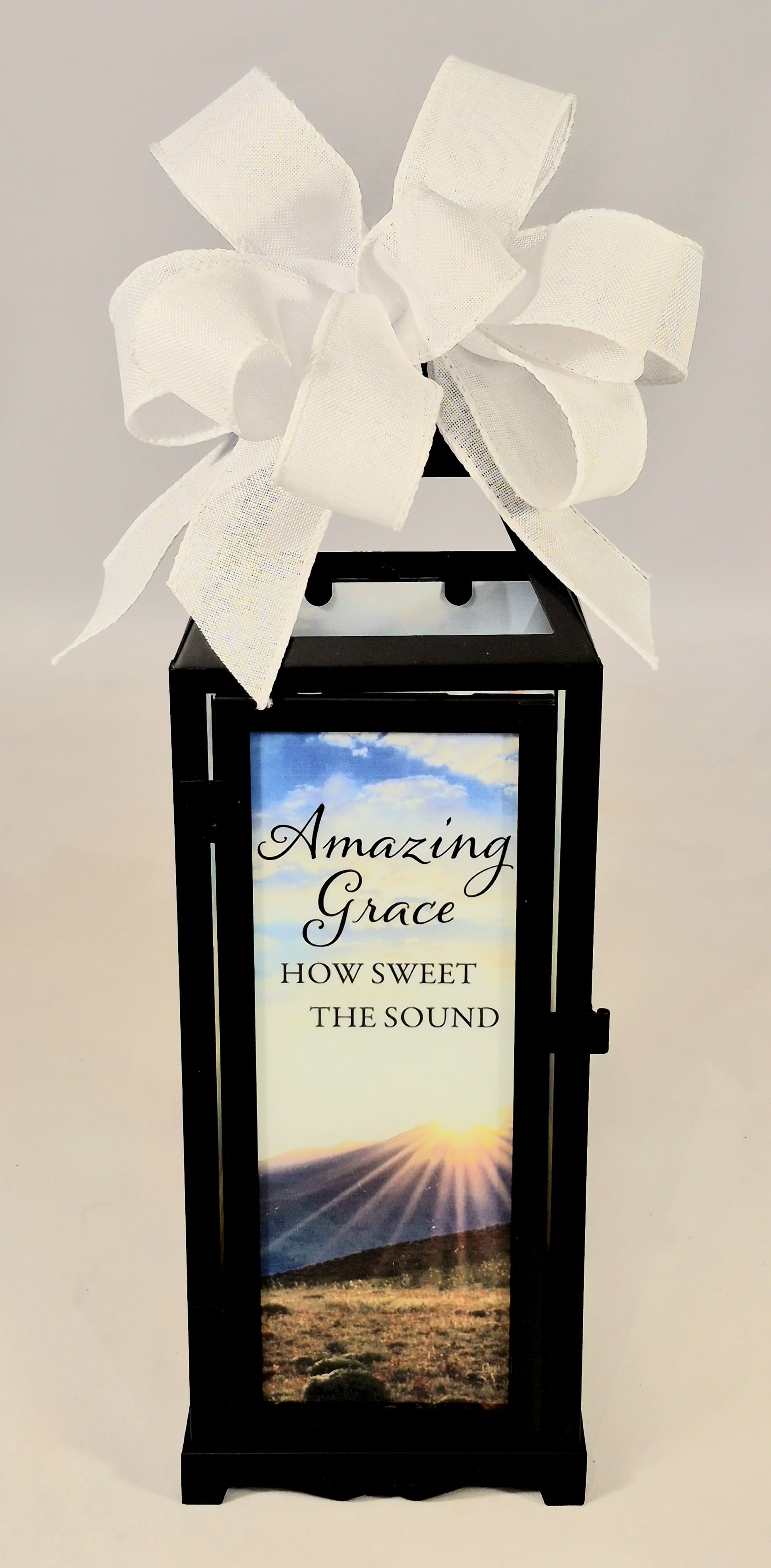 "Amazing Grace" Panoramic Lighted Lantern - A tall black metal lantern framed in glass, this lighted panoramic memorial piece greets the eye with a serene sunrise scene and the script "Amazing Grace How Sweet The Sound," making it an ideal sympathy arrangement or thoughtful Get Well or Thinking of You gift. The large white fabric bow at the handle softens the silhouette and lends a gentle, ceremonial feel-perfect for a funeral display, a bedside remembrance, or a quiet corner of the home where someone needs comfort. It also works well for seasonal moments like Easter or Passover without feeling limited to that holiday.  The lantern houses a built‑in flame-less LED candle with a 6‑hour ON / 18‑hour OFF timer and includes 3 AAA batteries, so it arrives ready to glow on its own schedule. Finished in powder‑coated black metal with clear glass panels, the piece measures approximately 4.75" wide x 15.5" high x 4.75" deep and can stand alone as a keepsake accent on a table, shelf, or memorial display. For someone grieving or recovering, this illuminated keepsake communicates care and remembrance in a peaceful, enduring way.  Choose deluxe if you would like to add a Silk arrangement on lid of lantern.