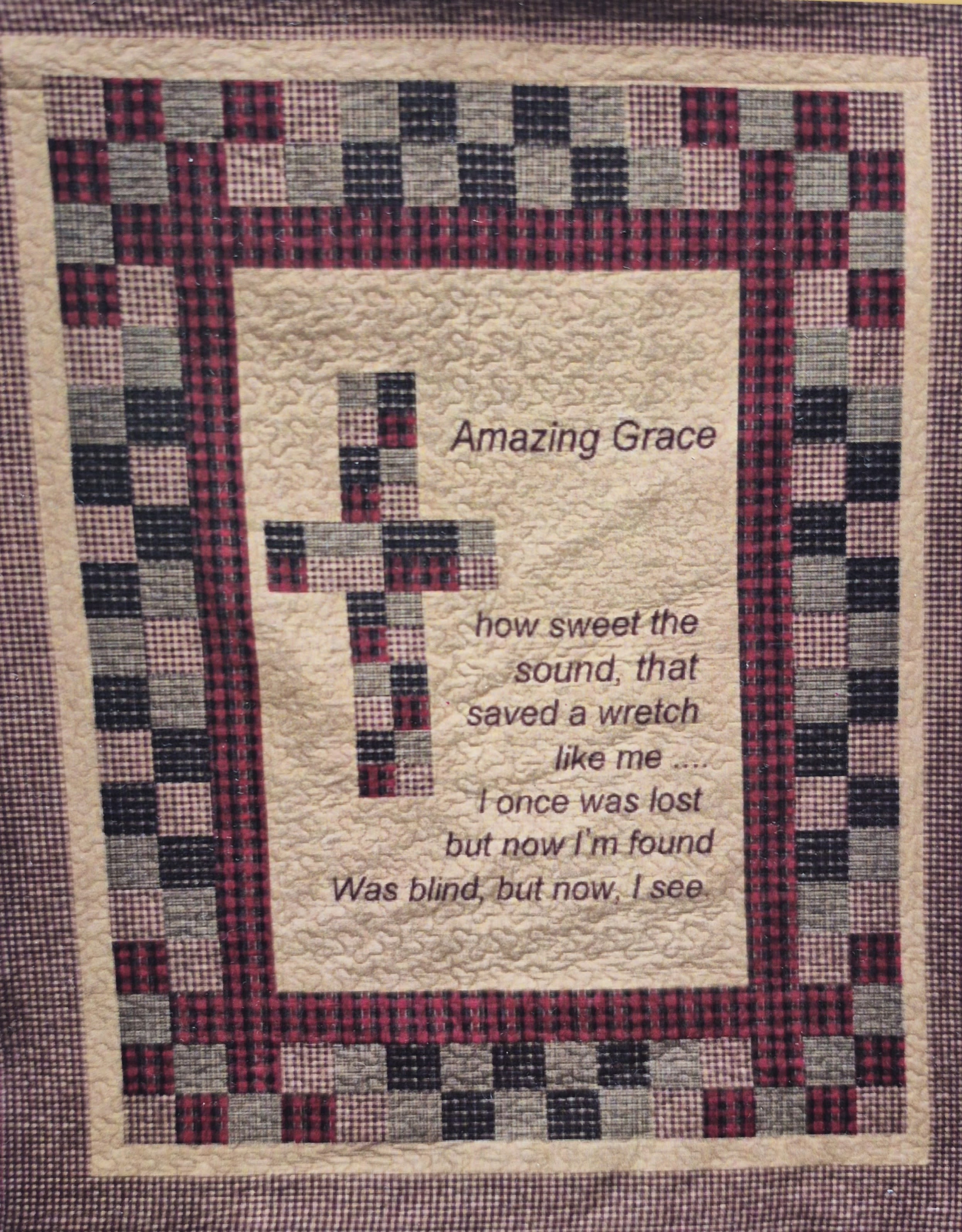 Amazing Grace Cross Quilted Throw  - A handcrafted quilted wall hanging centered with a patchwork cross in red, black and mixed plaid brings a quiet, devotional focus to any room-ideal as a sympathy or funeral gift or a comforting get-well surprise for someone who finds strength in faith. The embroidered lines of the hymn "Amazing Grace" on the cream textured field lend a reflective, hopeful tone so the person you're honoring feels seen and soothed.  Made from coordinated plaid and checked fabric blocks with textured quilting and a narrow outer frame, this 50 x 60 piece reads as warm, traditional home décor with a rustic, made-by-hand quality. Pair it with the slender black metal easel (available as a Deluxe add-on) for an upright presentation at a memorial service, beside a bedside, or in a prayer corner-the easel's simple tubular design keeps focus on the hanging while providing stable support.  Perfect for memorial settings, bedside comfort, housewarmings for faith-focused recipients, or as a gentle get-well gesture. Select the Deluxe option to include the easel for display; the easel is an extra charge of 15.00 choose Deluxe. 