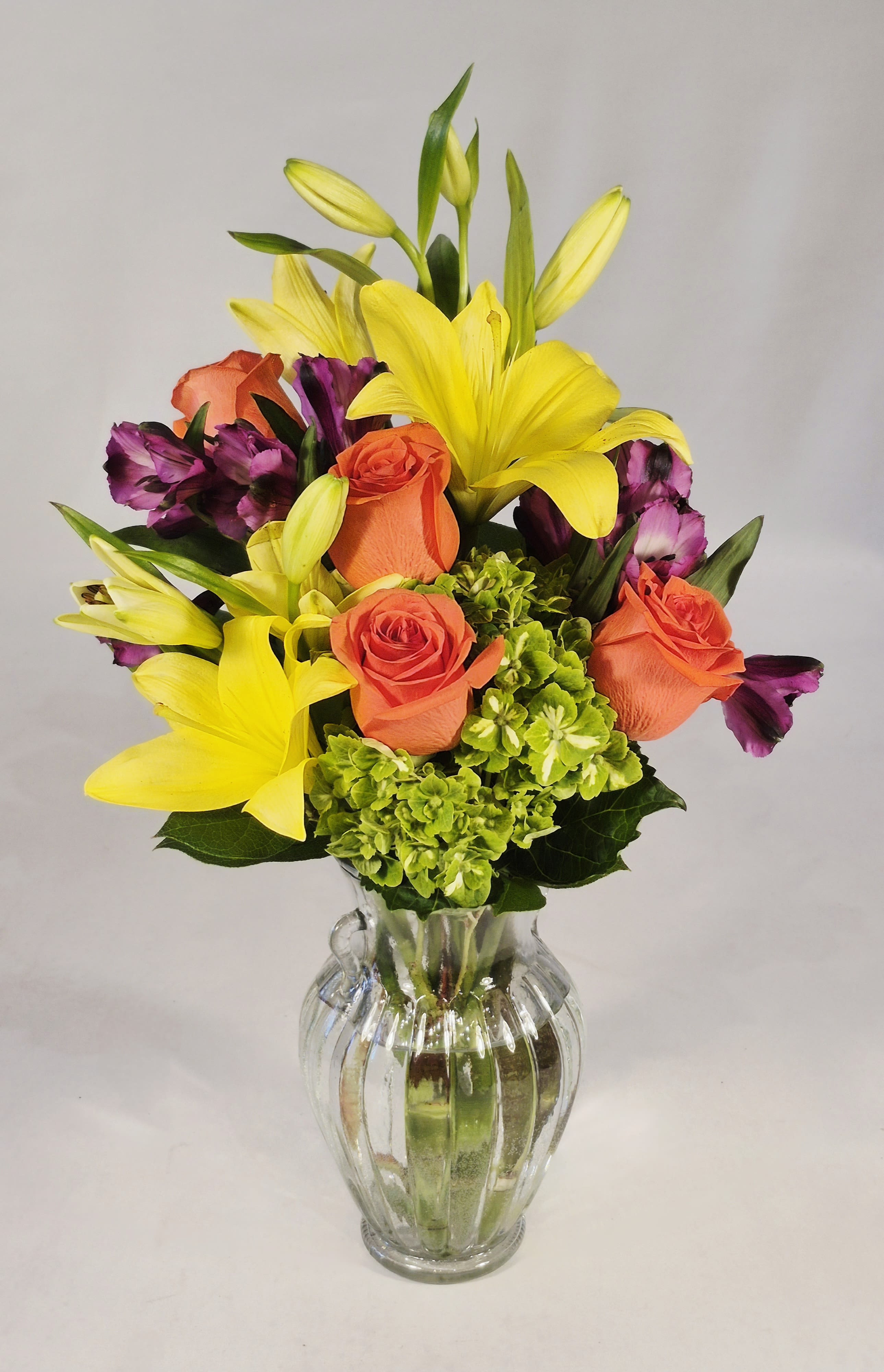 Bouquet of yellow lilies, orange roses, and purple flowers in a clear glass vase